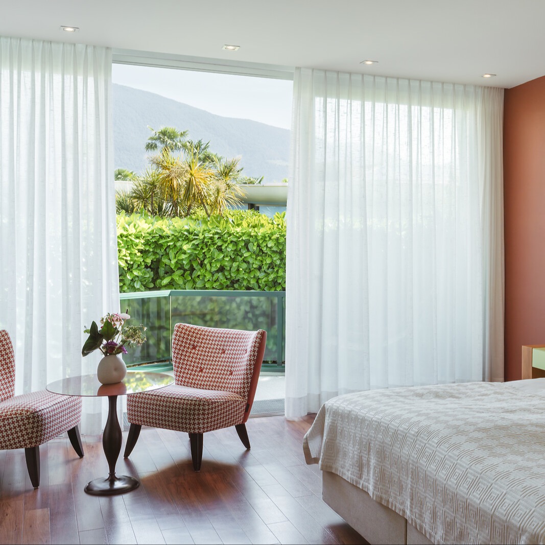 Modernes Schlafzimmer mit weissen Vorhängen, gemustertem Sessel, Blick auf Balkon mit Palmen und Berglandschaft.