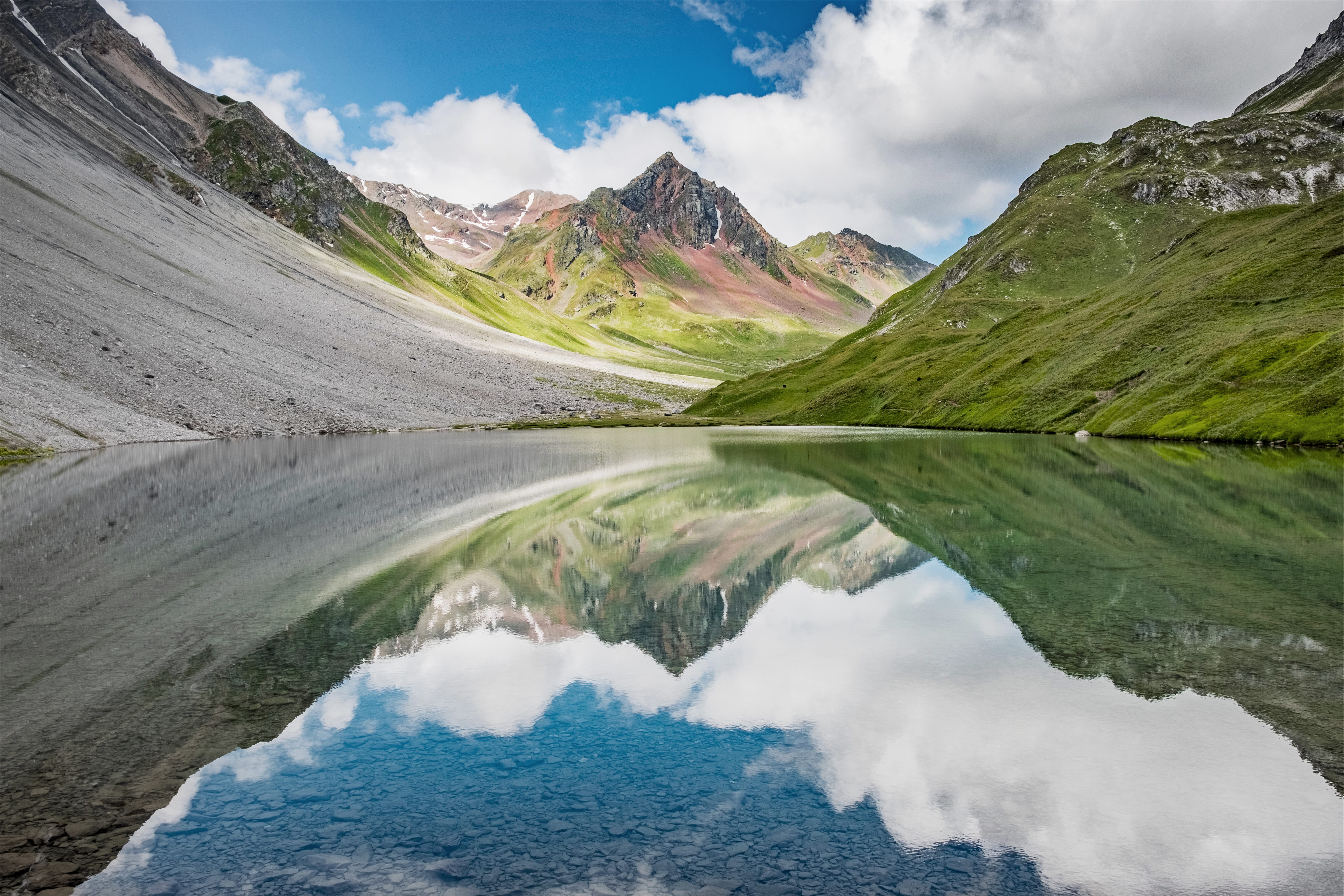 Mountain valley with green slopes and rocky peaks reflected in a calm alpine lake under a blue sky with white clouds.