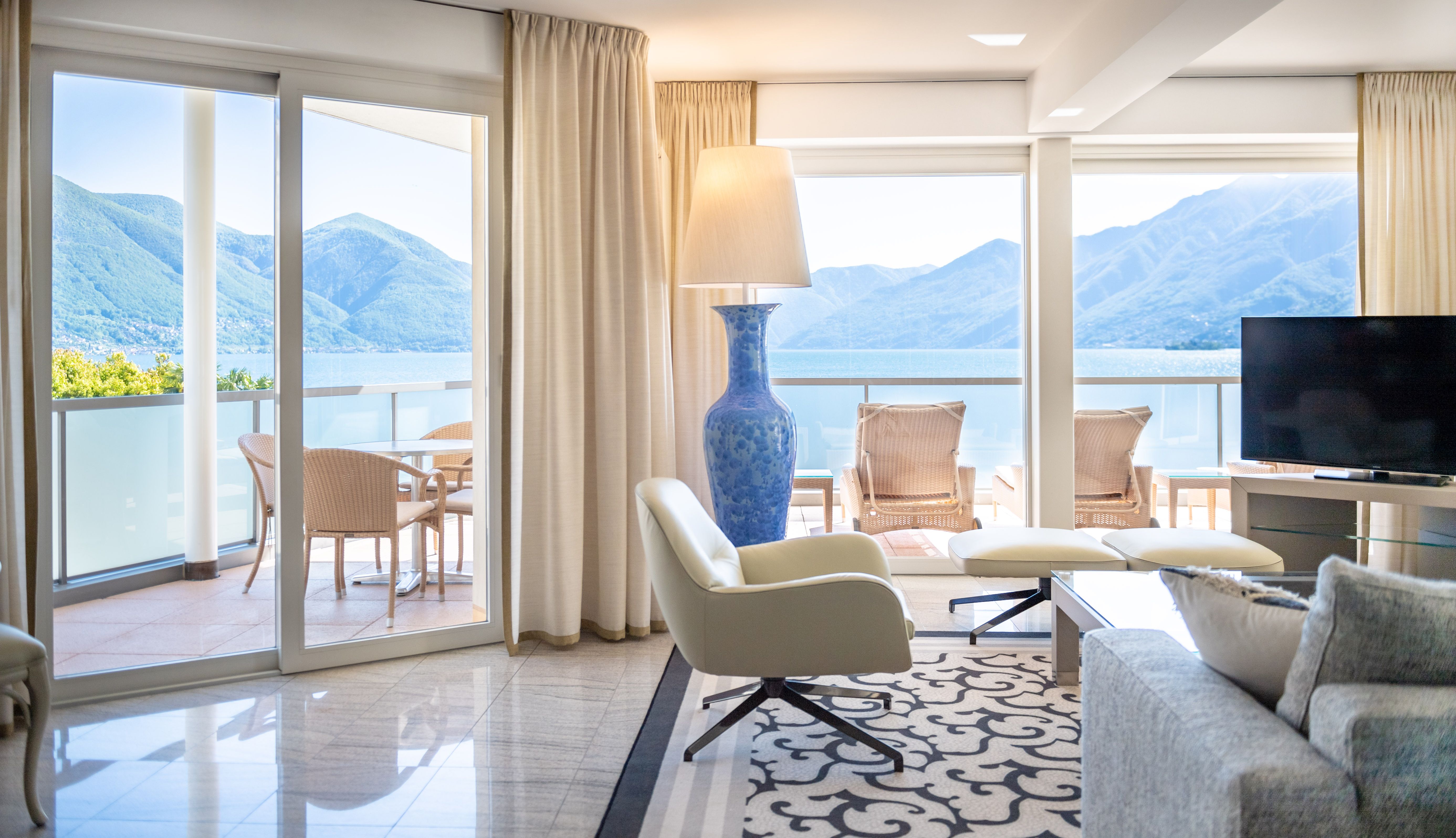 Modern hotel suite with floor-to-ceiling windows overlooking a lake and mountains, featuring neutral furnishings and a balcony.
