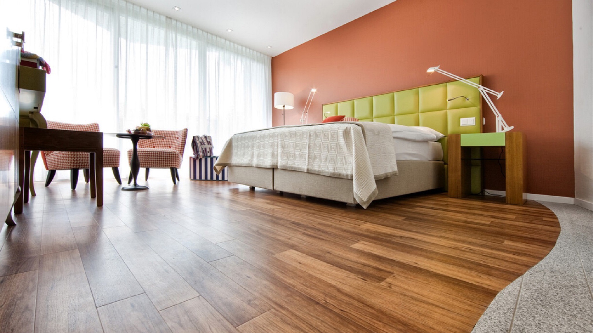 Spacious double room with orange accent wall, green upholstered bed, two patterned armchairs, natural light, and stylish details.