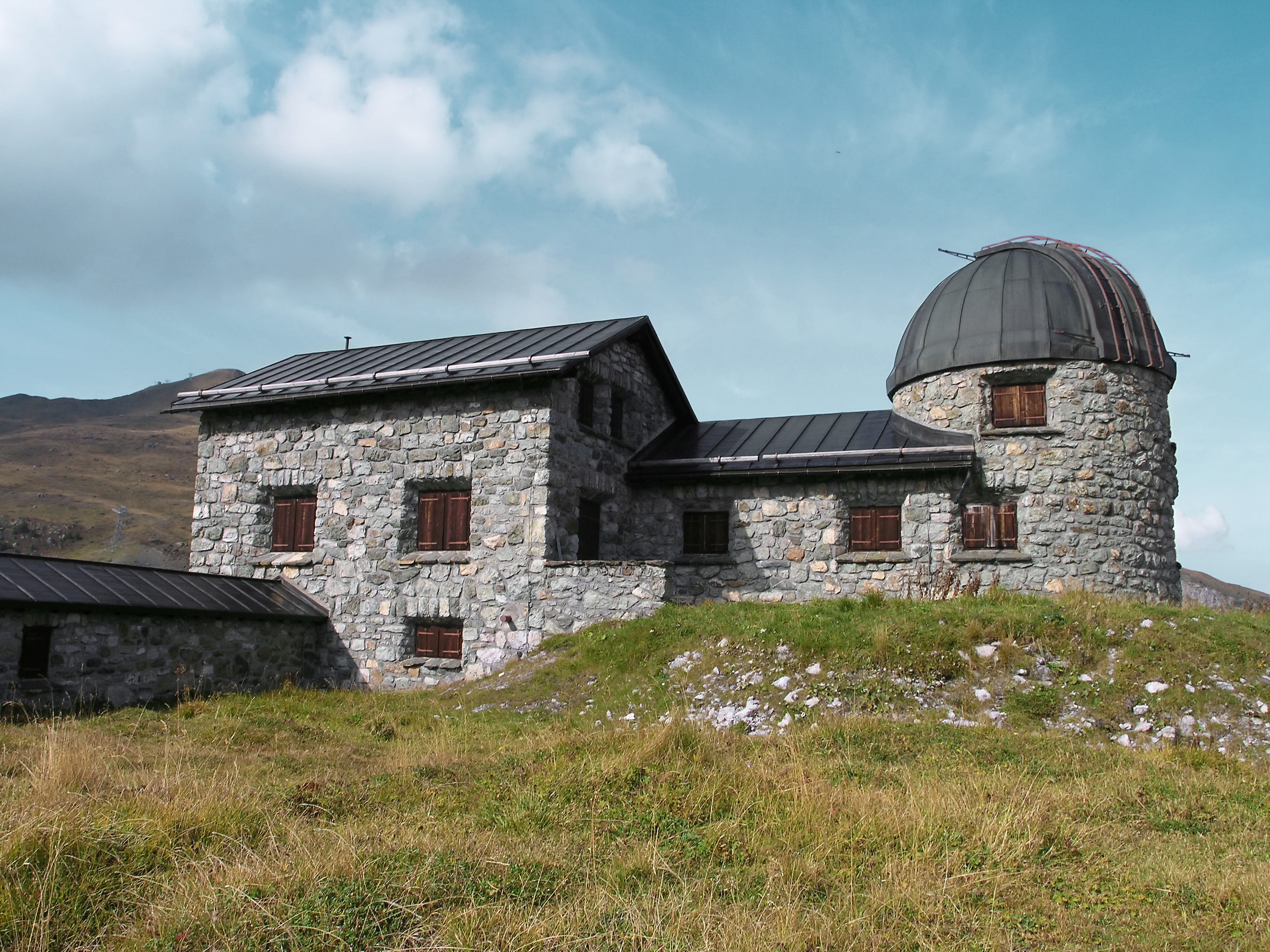 Stone observatory building with domed tower on grassy hillside under blue sky with white clouds and mountain backdrop.