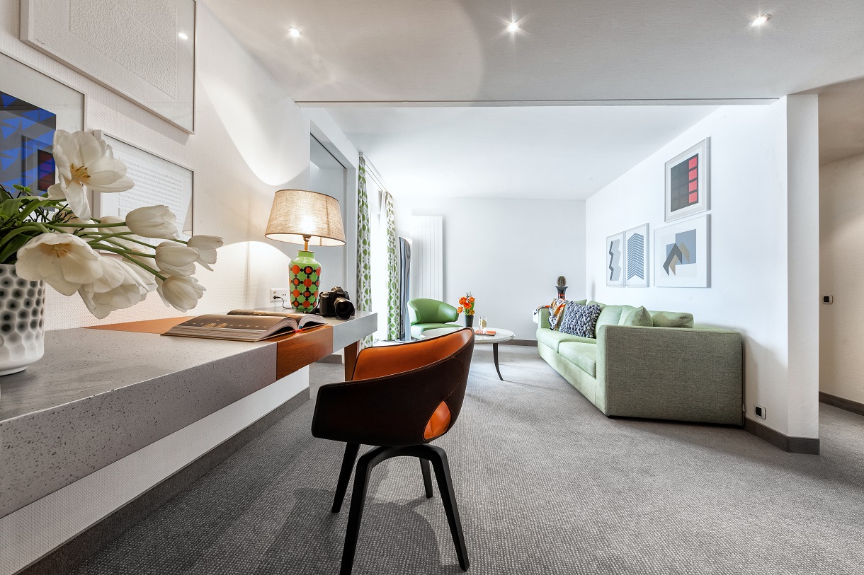 Stilvoll eingerichtetes Wohnzimmer mit hellgrüner Couch, Design-Sesseln, Schreibtisch mit Tulpenvase und Lampe, modernem Teppich und dekorativer Wandkunst.