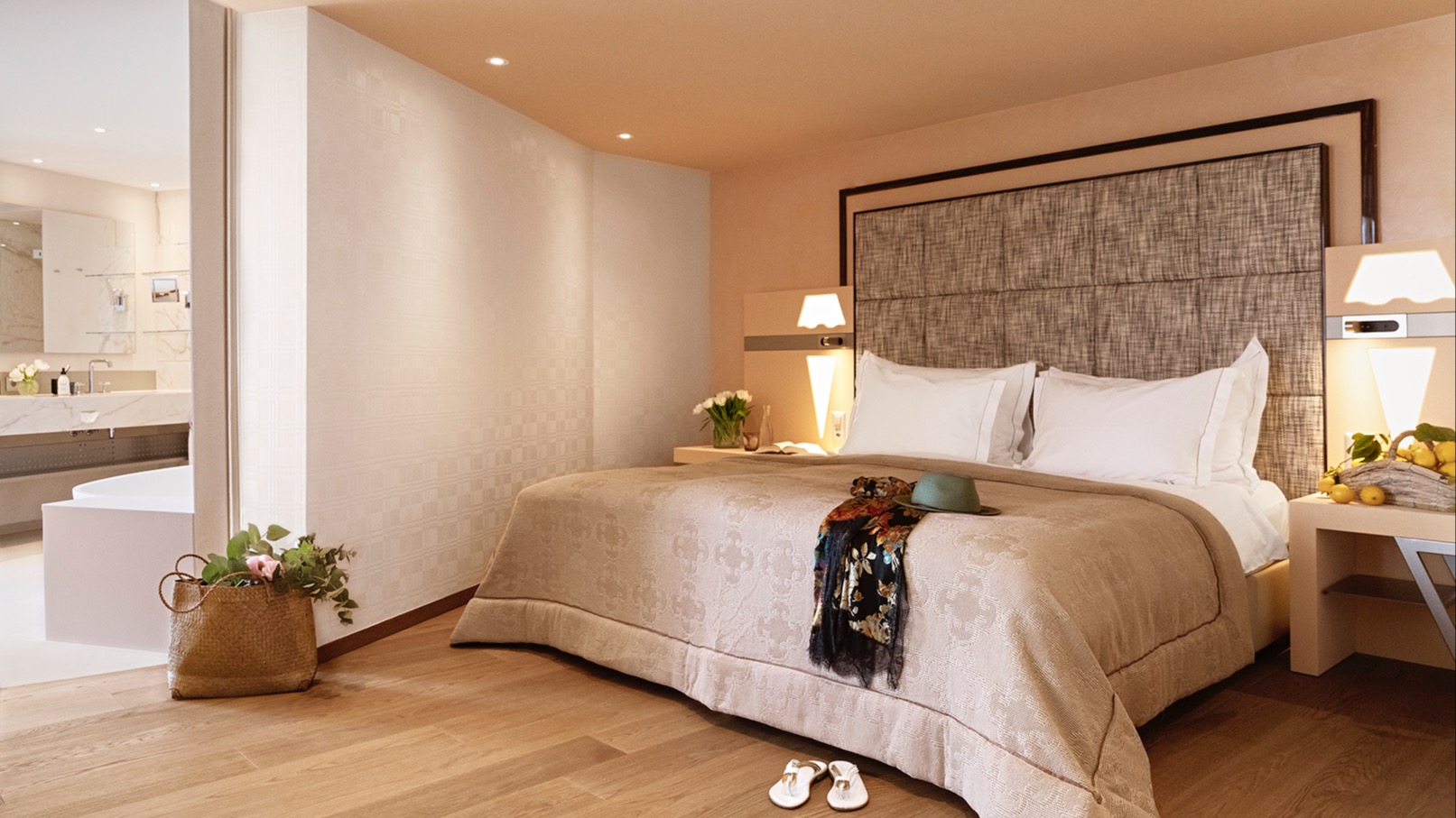 Upscale hotel suite with king-size bed, floral accents, open connection to bathroom with freestanding bathtub, wooden flooring, and accessories.