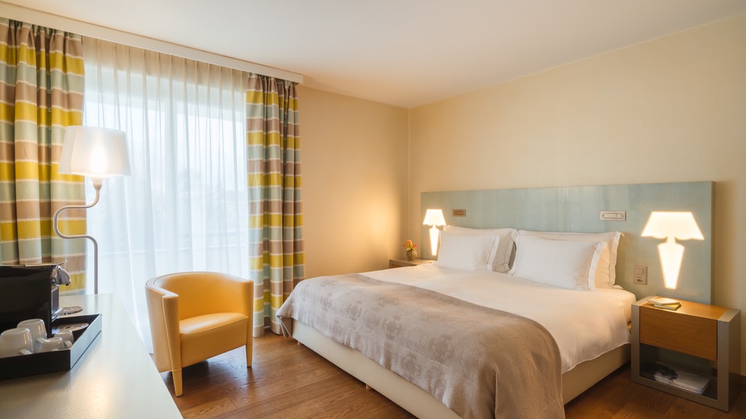 Freundliches Hotelzimmer mit großem Doppelbett, hellblauem Kopfteil, Parkettboden, gelbem Designersessel, Stehlampe und farbenfrohen Vorhängen mit Streifenmuster.