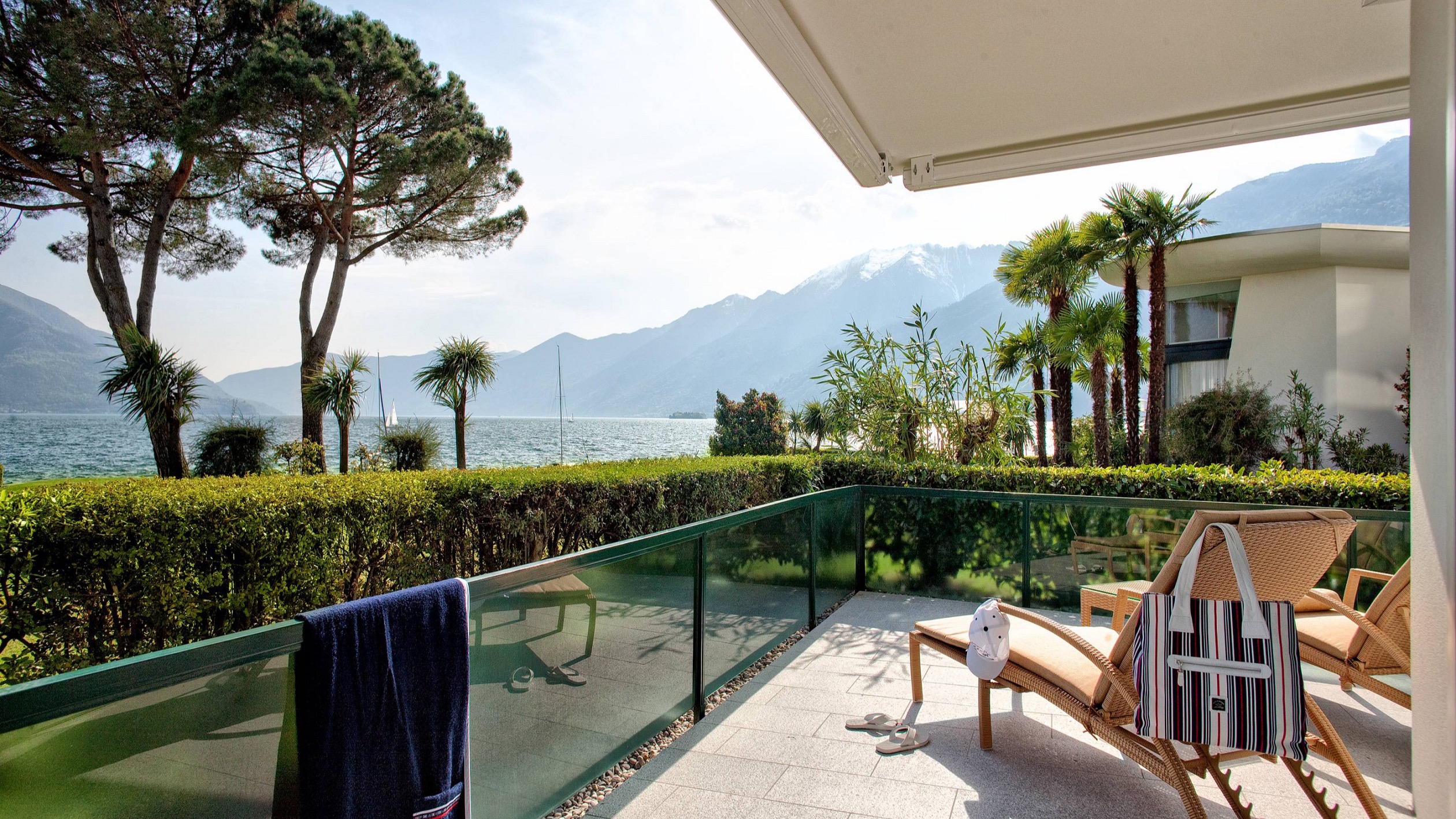 Private Terrasse mit Sonnenliegen, Badetasche, Blick auf Palmen, den See und die umliegenden Alpen.