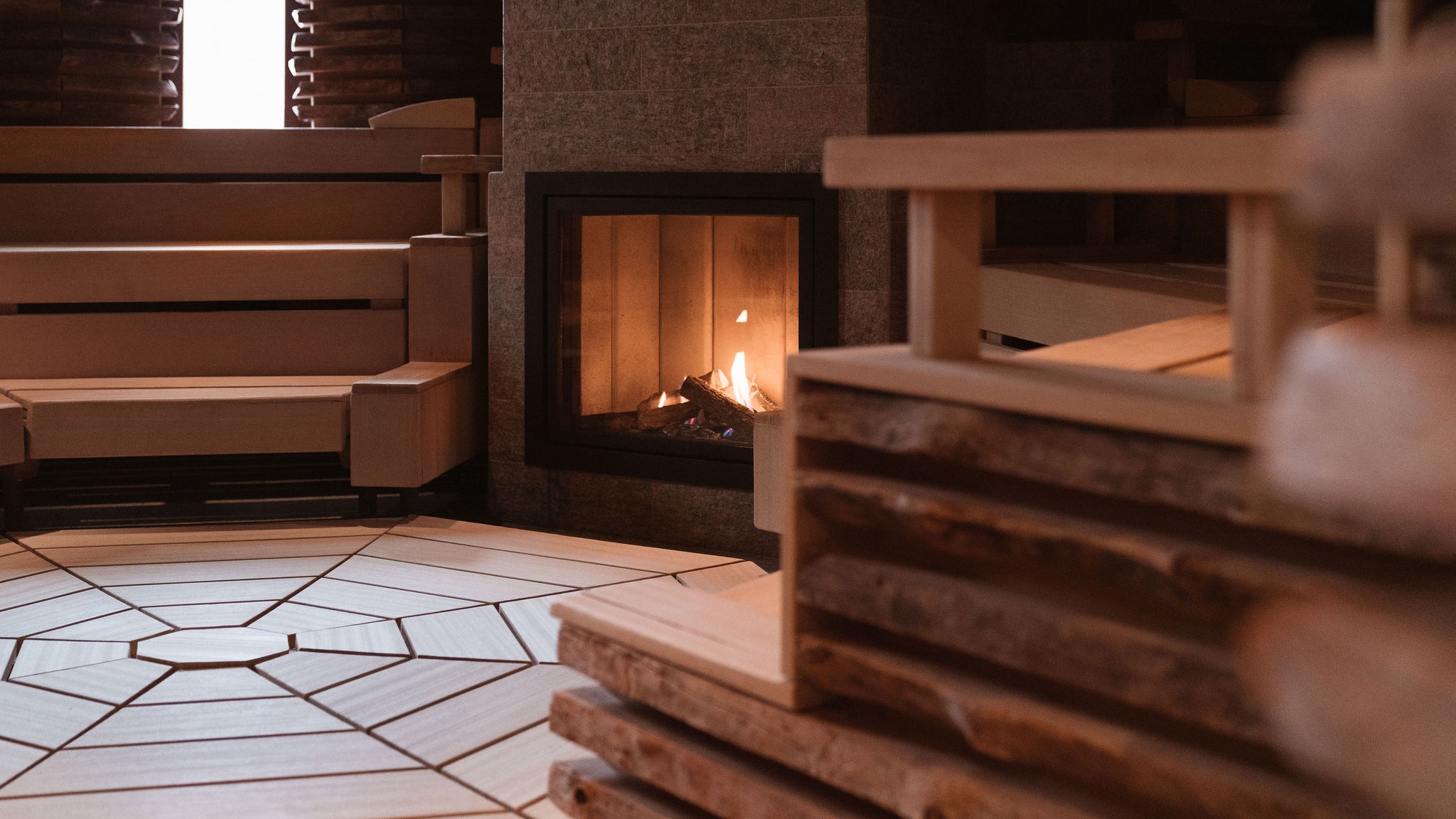Gemütliches Sauna-Interieur mit Holzbänken, weißem Fliesenboden und einem glühenden Kamin, dessen Flammen durch das Glas sichtbar sind.