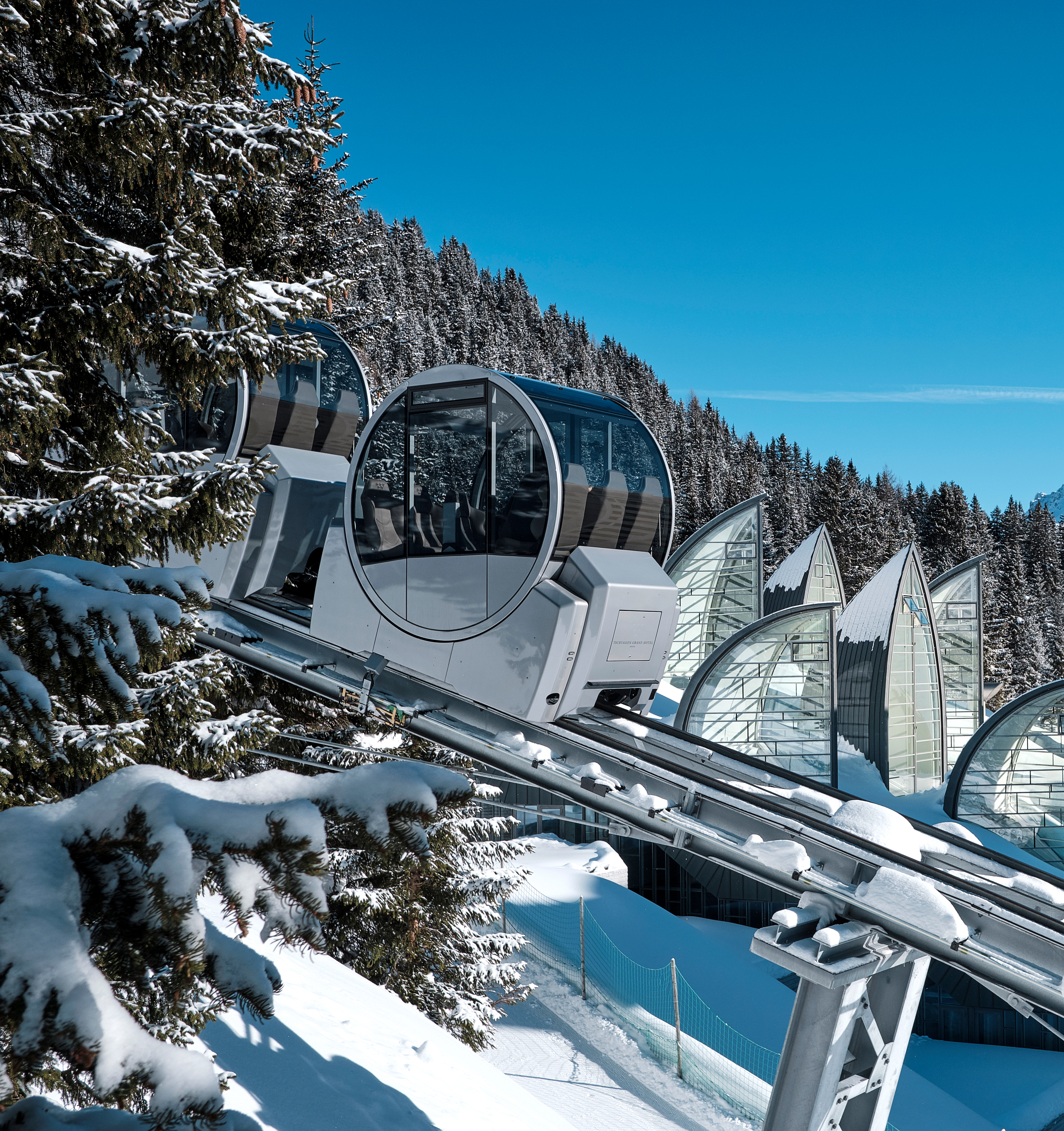 Moderne Seilbahngondeln mit Panoramafenstern fahren über verschneite Tannen vor blauem Himmel in den Alpen.