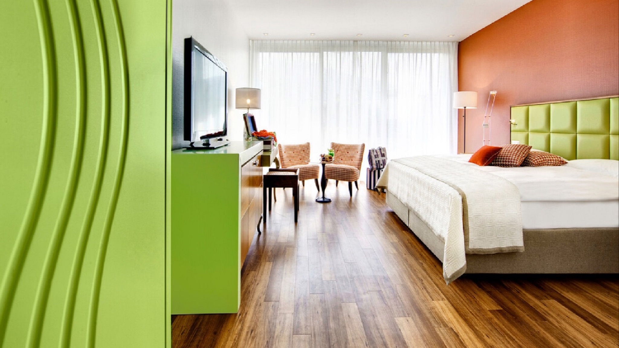 Farbintensives Hotelzimmer mit grünem Schrank, orangefarbener Wand, gelb-grünem Kopfteil, Parkettboden und Panoramafenster mit Sitzbereich.