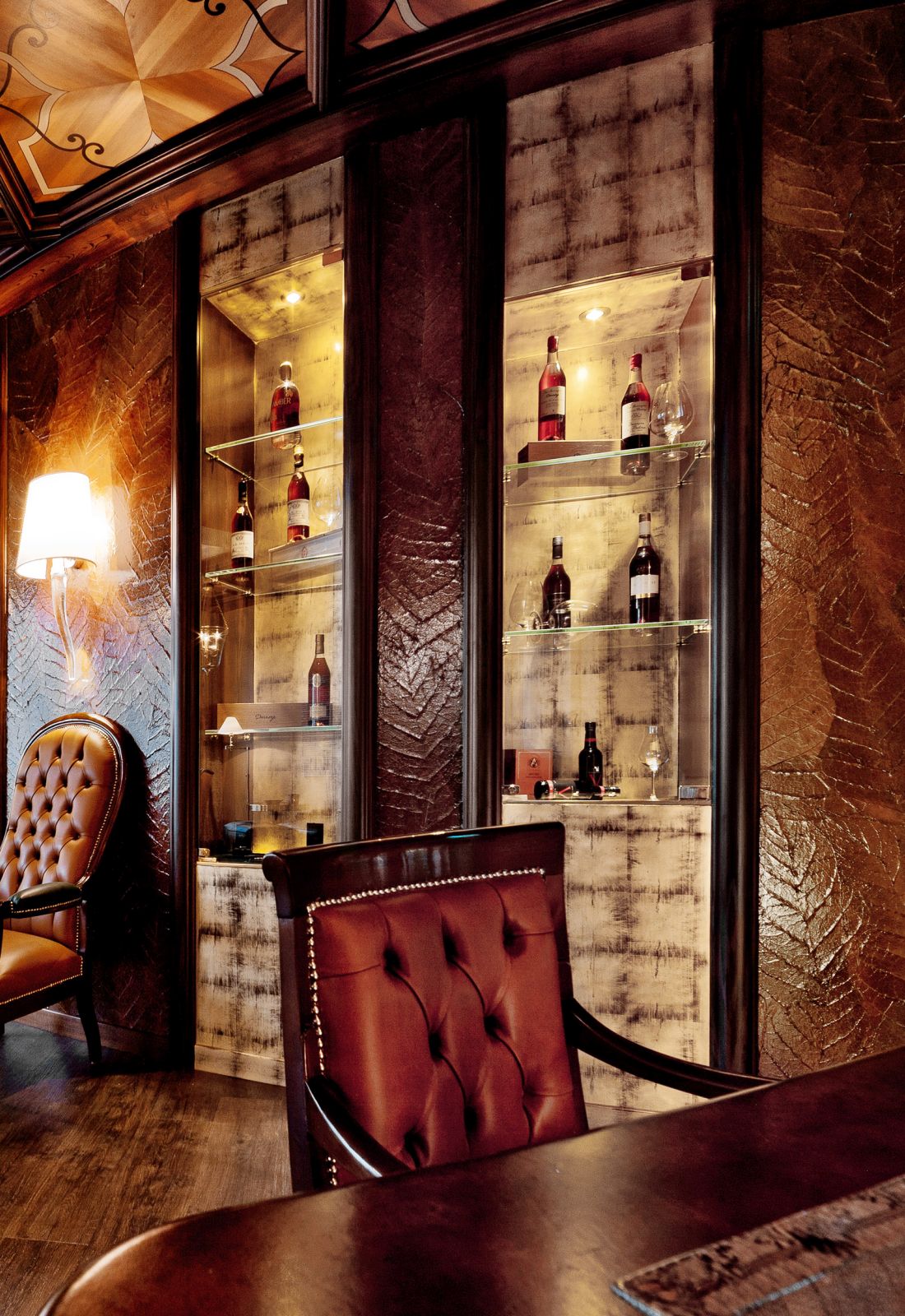 Elegant wine display cabinets with glass shelves and interior lighting, featuring wine bottles, flanked by burgundy Lounge chairs.