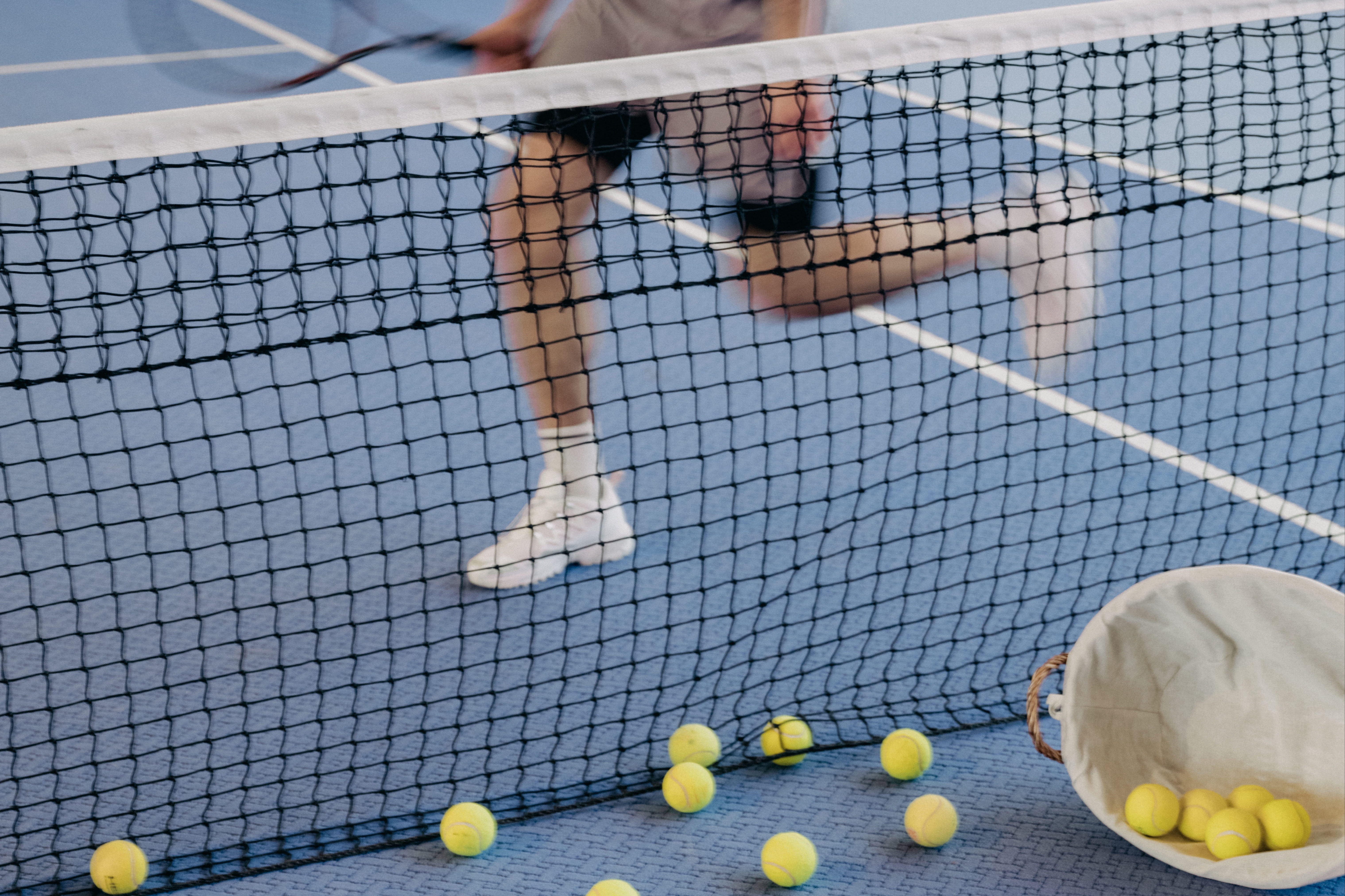 Tennisnetz auf blauem Platz mit gelben Bällen und verschwommenen Spielern im Hintergrund