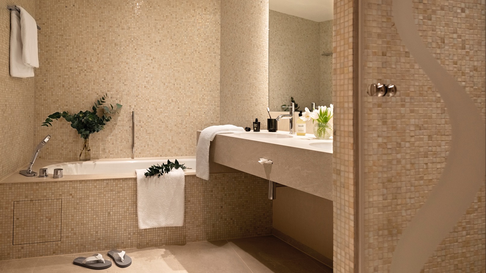 Elegantes Badezimmer mit beigen Mosaikfliesen, Einbaubadewanne, großem Spiegel, Doppelwaschbecken, weißen Handtüchern und Eukalyptusdeko.
