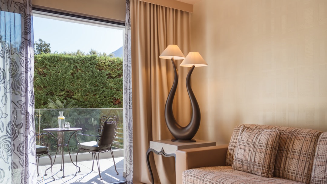 Elegant living room with beige sofa, artistic designer lamp, glass door leading to a balcony with wrought iron bistro set, floral curtains, and garden view.