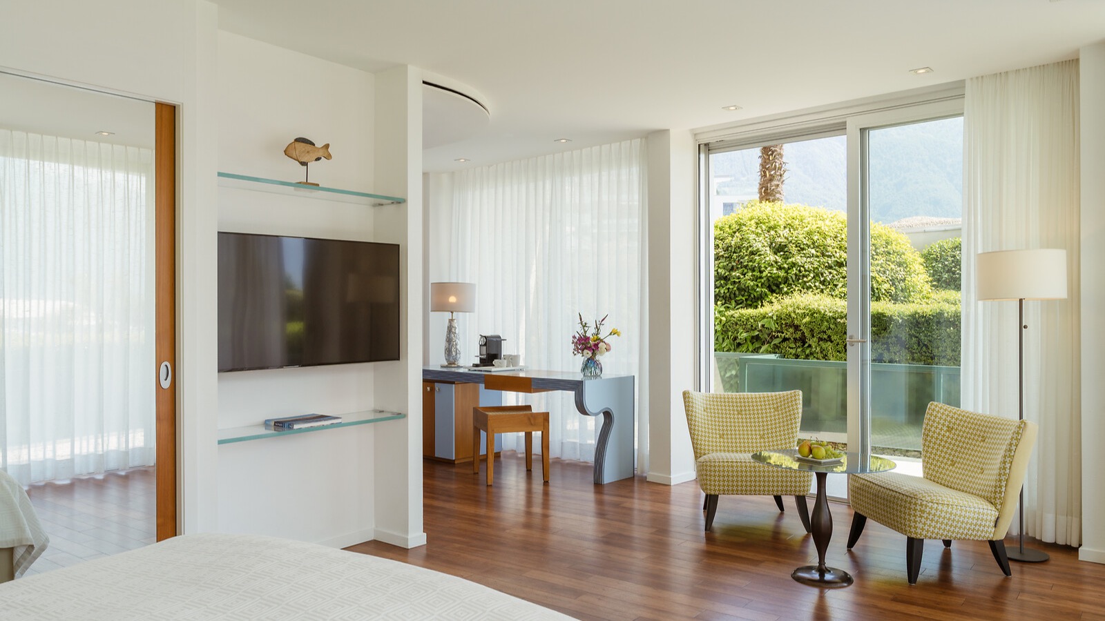 Modernes Hotelzimmer mit Flachbildfernseher, Schreibtisch, gelben Sesseln und Balkonzugang mit Gartenblick.