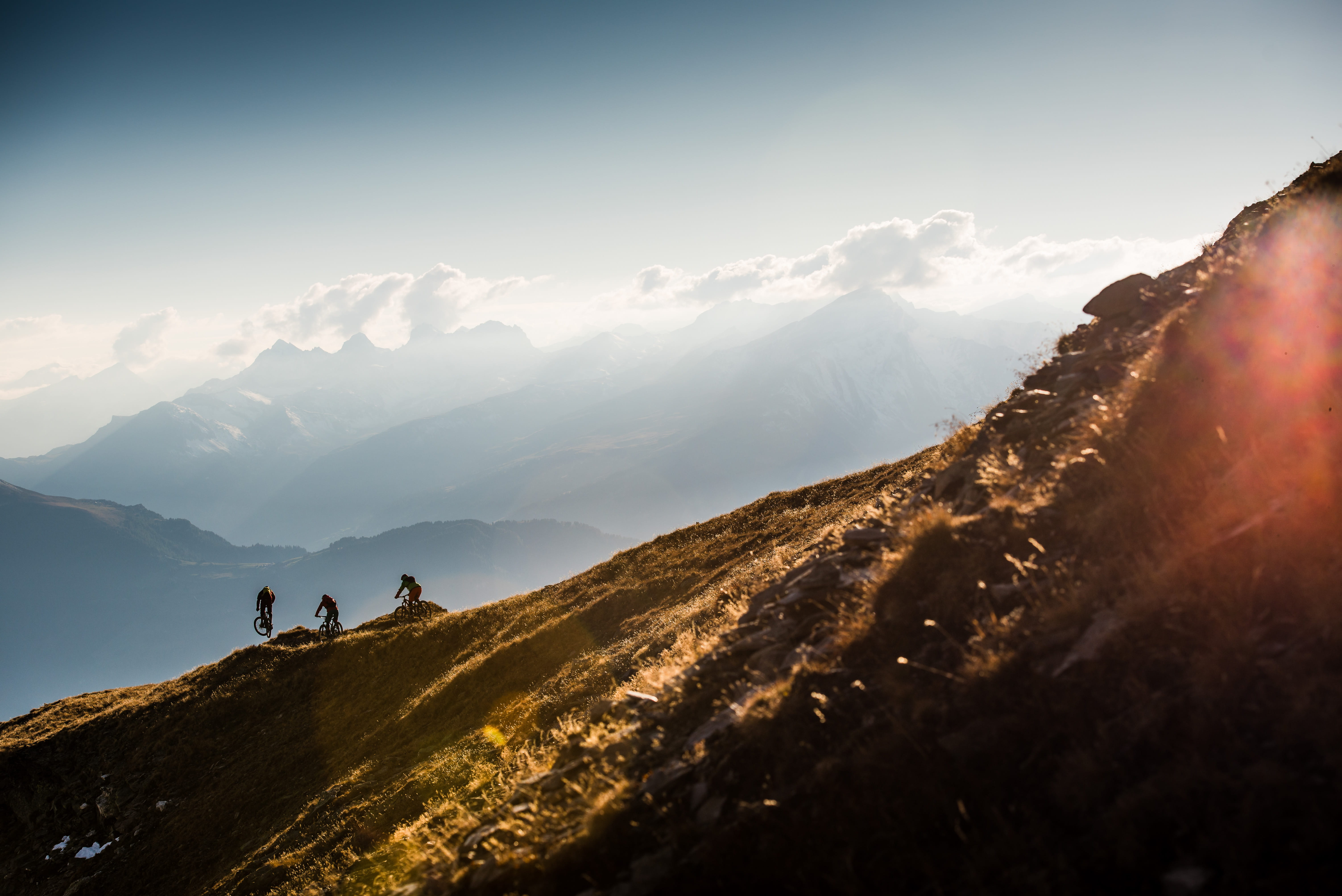 Drei Mountainbiker fahren bei Sonnenuntergang einen Berghang hinab, Alpenpanorama mit Wolken im Hintergrund.