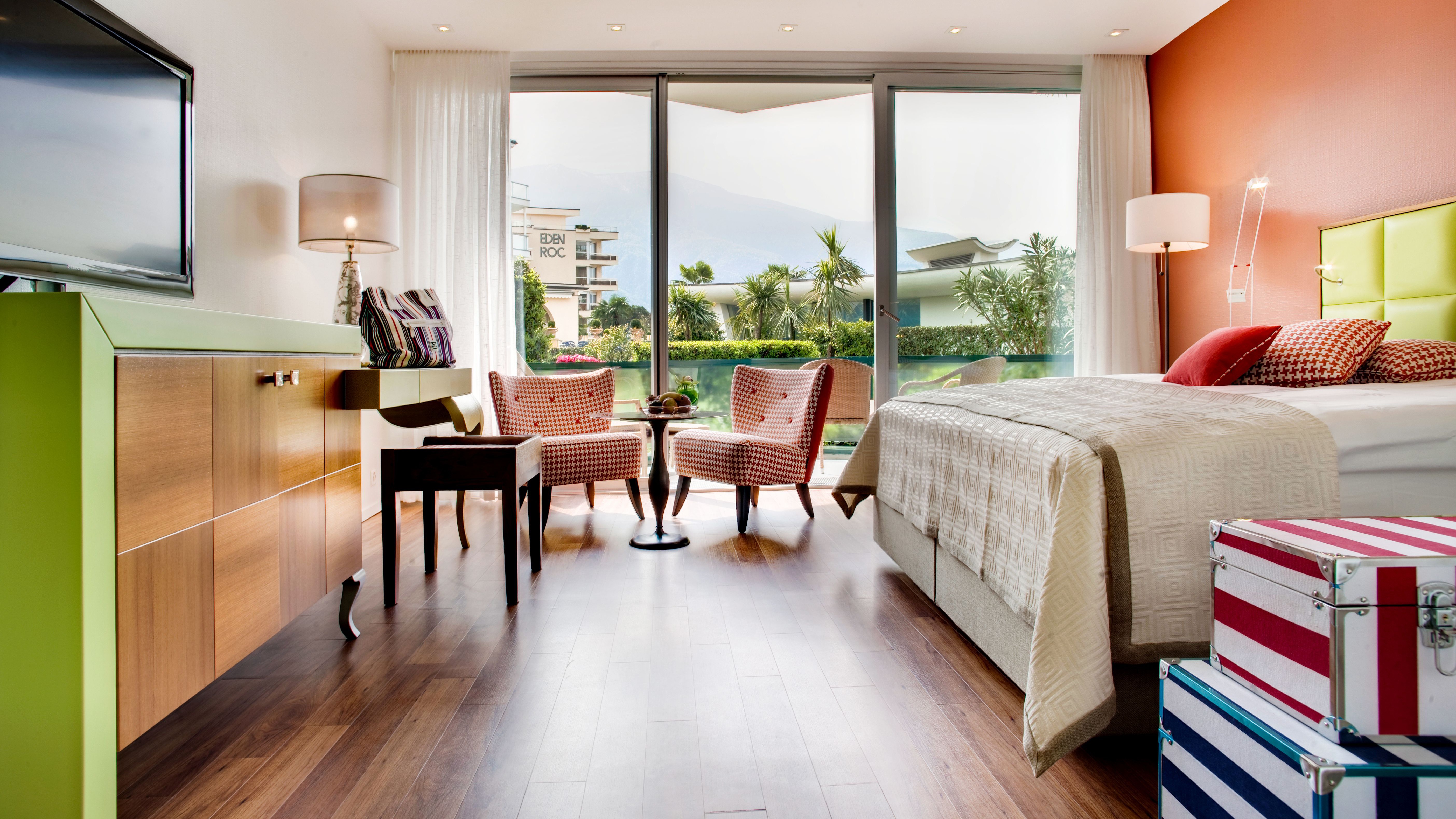 Color-accented hotel room with green sideboard, two red armchairs, garden access, wooden floor, and decorative trunks.