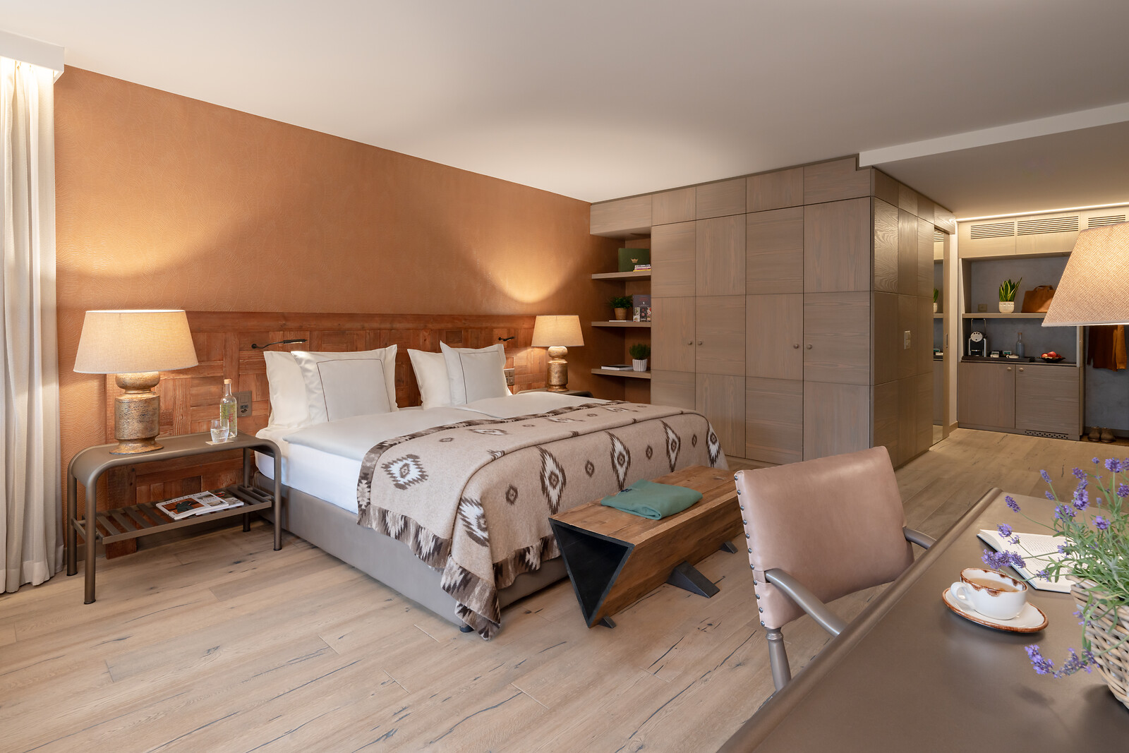 Modernes Junior Suite Zimmer mit Doppelbett, warmen Holz- und Erdtönen, Sitzbereich und integrierter Küchenzeile im Hintergrund.