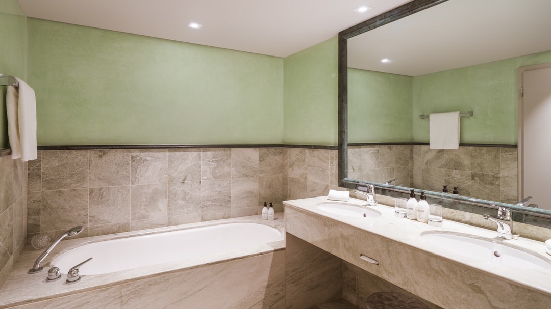 Elegant bathroom with green painted walls, large mirrors, marble tiles, built-in bathtub, and double vanity with toiletries.