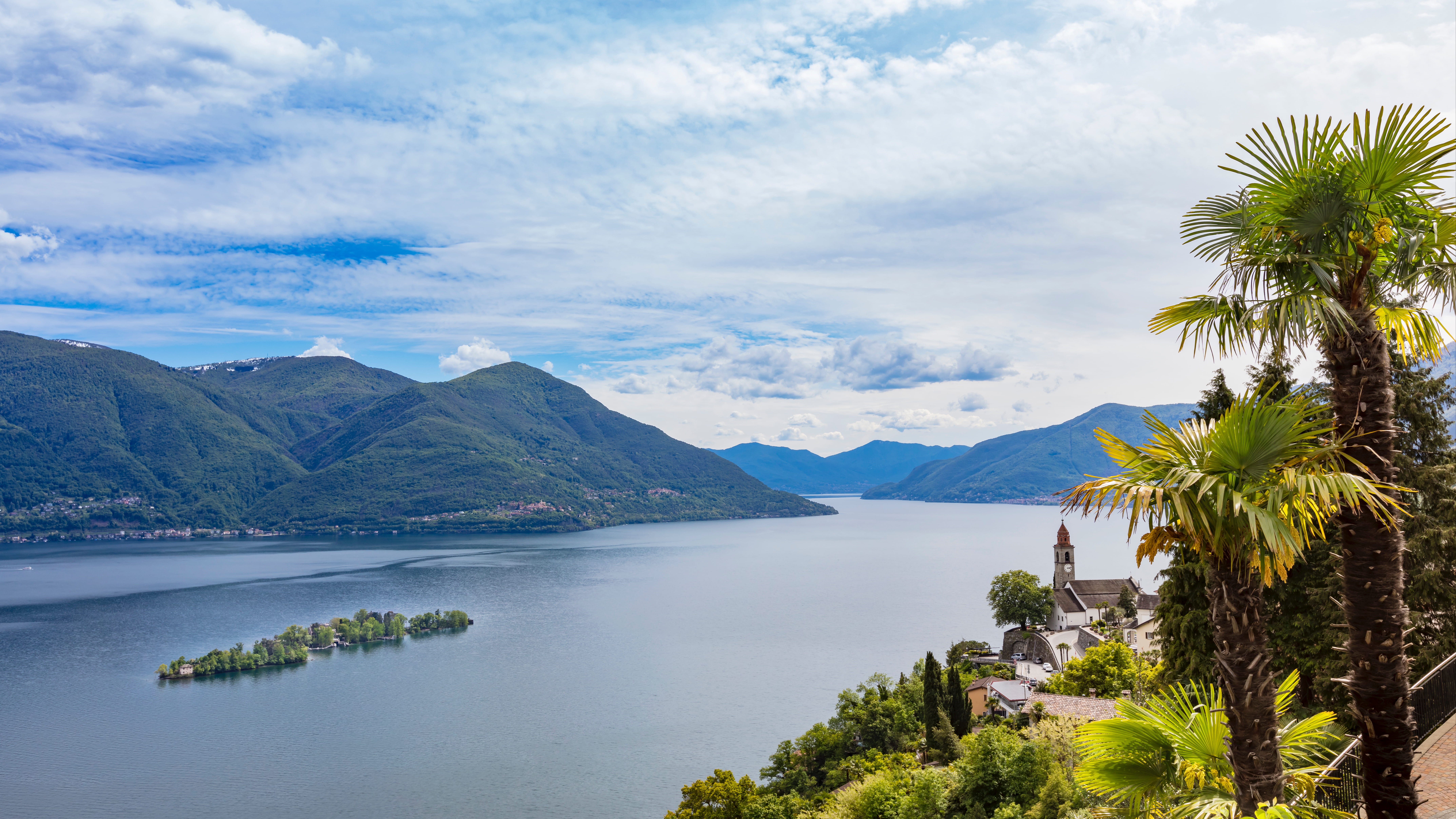 Panoramablick vom Ronco sopra Ascona über den Lago Maggiore mit Kirche, Palmen, bewaldeten Bergen und einer kleinen Insel im See.