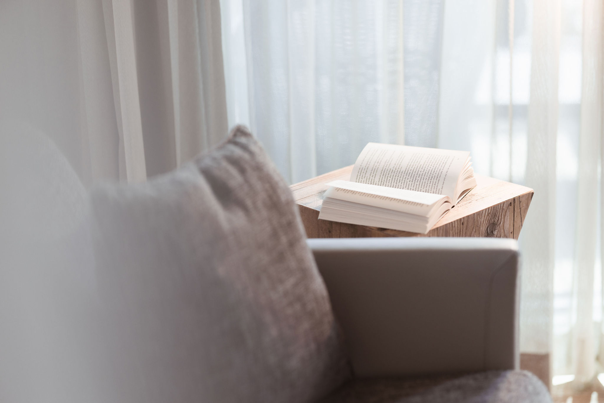 Open book resting on wooden tray beside gray cushion with white sheer curtains and bright window in background.