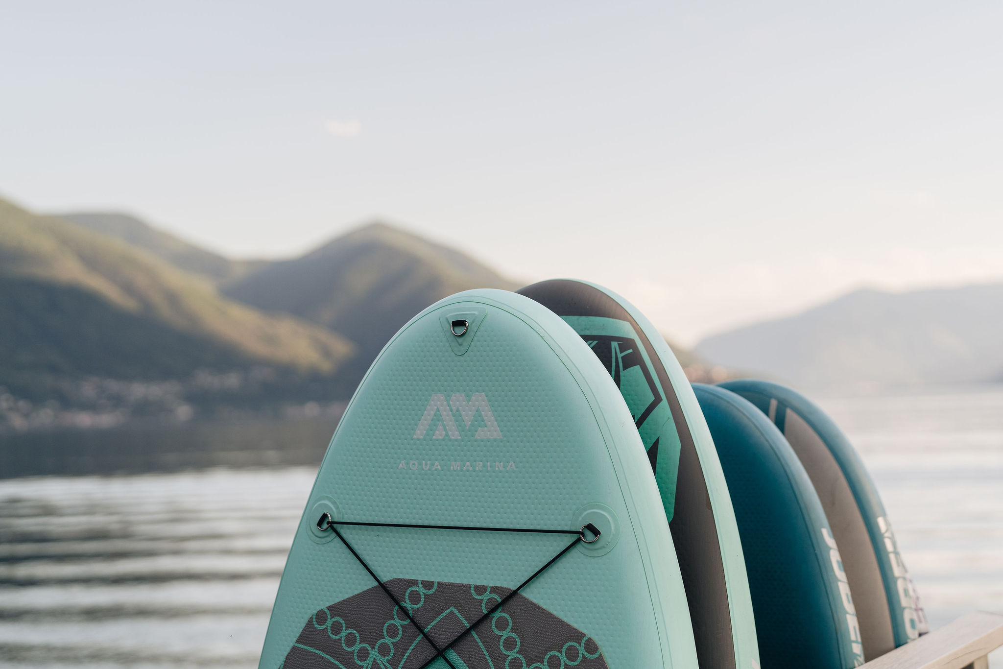 Türkisfarbene Aqua-Marina-Paddleboards, die auf einem Steg gestapelt sind, mit verschwommenen Bergen und Wasser im Hintergrund.