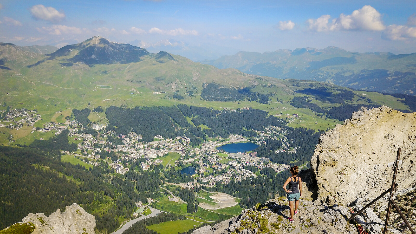 Hiker standing on rocky mountain peak overlooking alpine valley with village, blue lakes, green forests, and distant mountains.