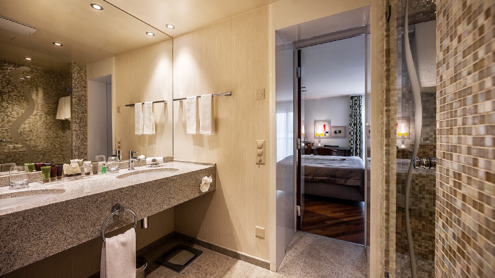 Luxury hotel bathroom with large mirror, double sink, walk-in mosaic-tiled shower, and view into the elegant bedroom.
