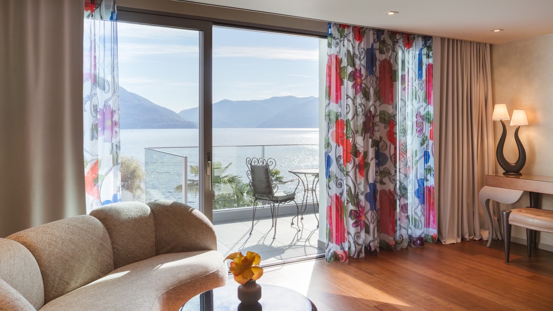 Bright living room with panoramic window, floral patterned curtains, beige designer sofa, glass table with yellow flower, and balcony overlooking lake and mountains.