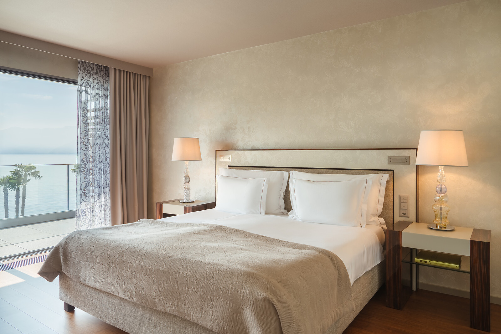 Luxurious bedroom with double bed, beige bedding, bedside tables with lamps, floor-to-ceiling windows, and lake view with palm trees.
