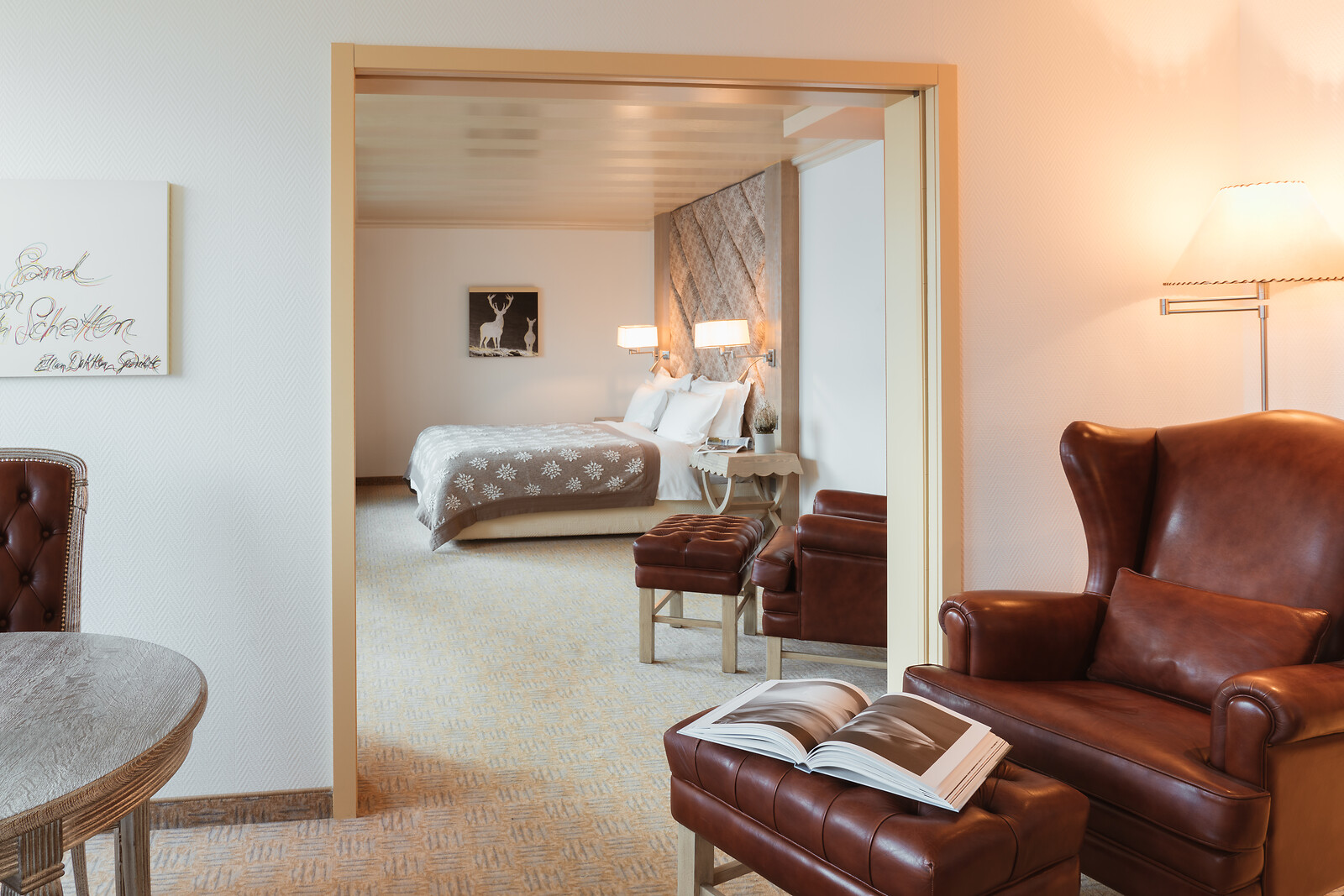 Elegant hotel suite with a brown leather wingback chair and footstool in the foreground, leading through sliding doors to a spacious bedroom with a large double bed, woven feature headboard and soft neutral furnishings.
