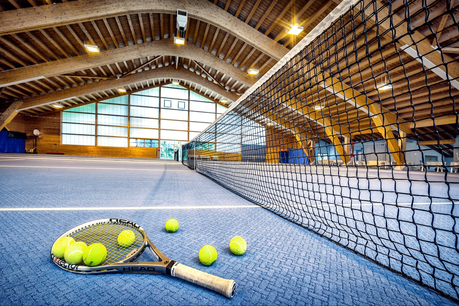 Indoor tennis court with arched wooden ceiling, net, and tennis racket with yellow balls on blue court surface.