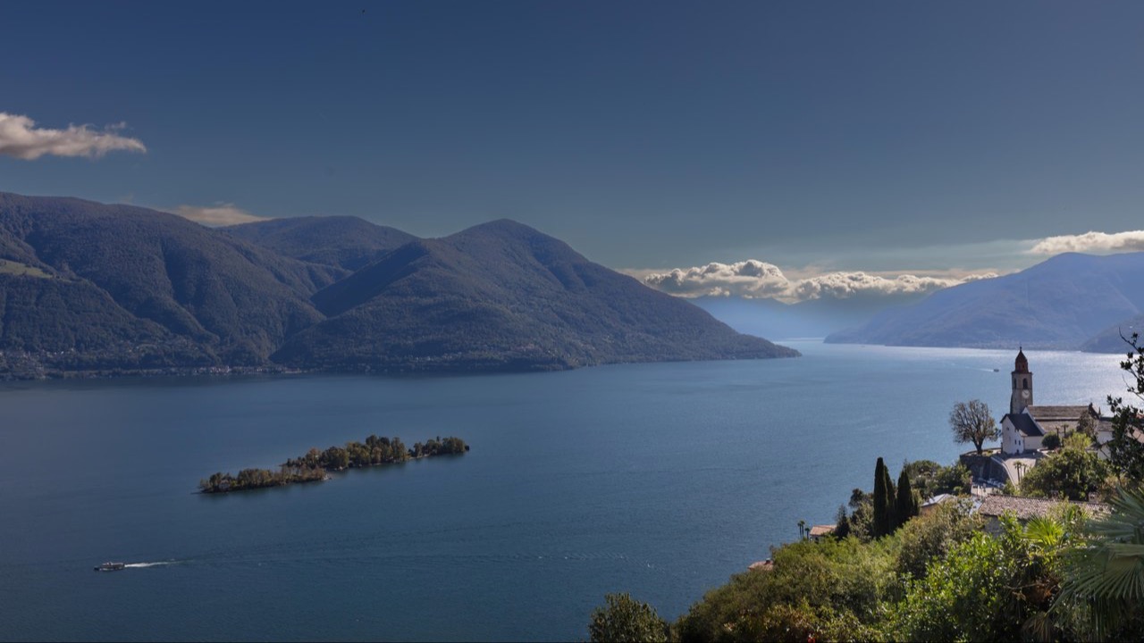 Panoramic view over Lake Maggiore with forested mountains, a small island in the lake, and a church on a hillside by the shore.