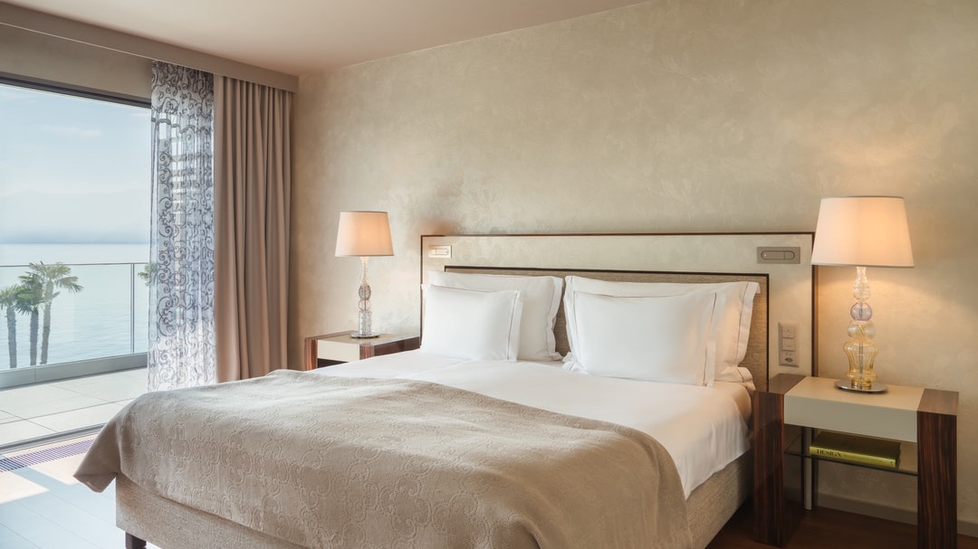 Luxuriöses Schlafzimmer mit Doppelbett, beigen Betttextilien, Nachttischen mit Lampen, bodentiefen Fenstern und Seeblick mit Palmen.