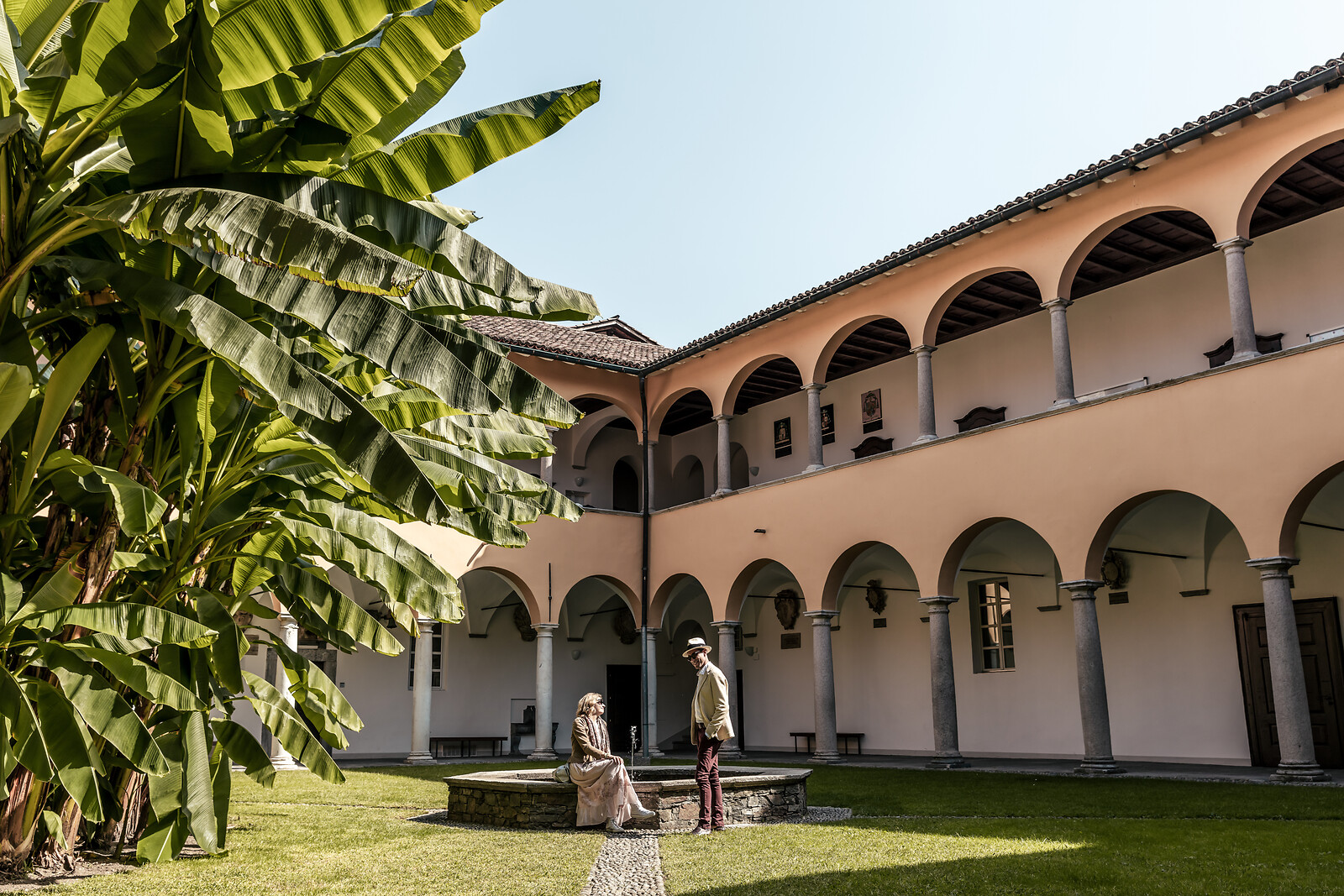 Two people in period costume stand in a courtyard with arched colonnade building and tall banana plants under blue sky.