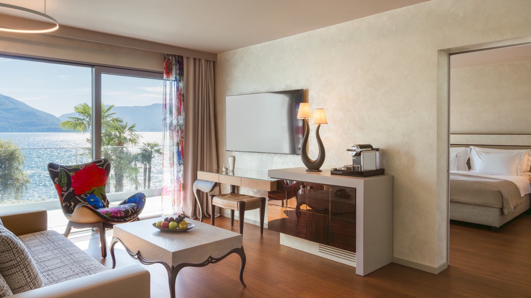 Wohnzimmer mit Panoramafenstern, Blick auf See und Palmen, modernem Mobiliar, Flachbildfernseher, Designlampe, Kaffeestation und Zugang zum Schlafzimmer.