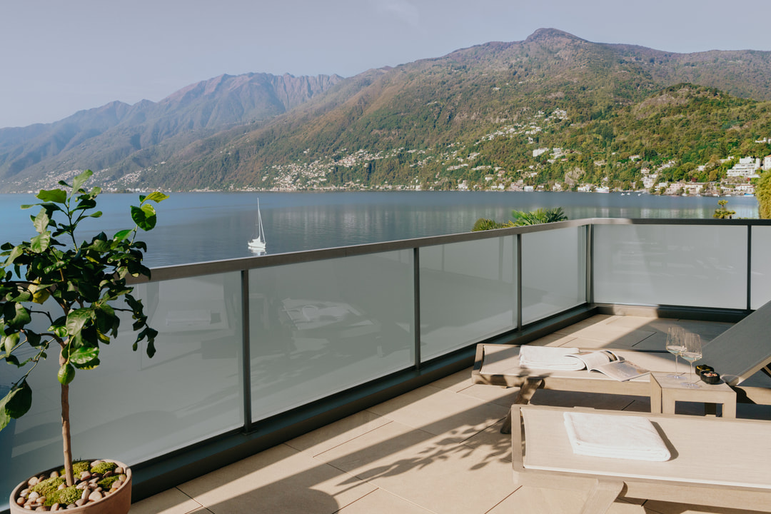 Moderne Terrasse mit Glasgeländer, Liegestühlen und Panoramablick auf einen See mit Bergen und Segelboot im Hintergrund.