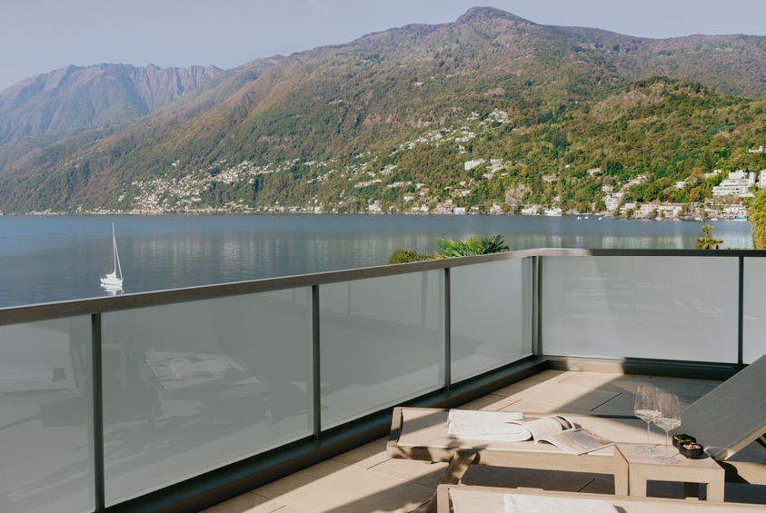 Moderne Terrasse mit Glasgeländer, Liegestühlen und Panoramablick auf einen See mit Bergen und Segelboot im Hintergrund.