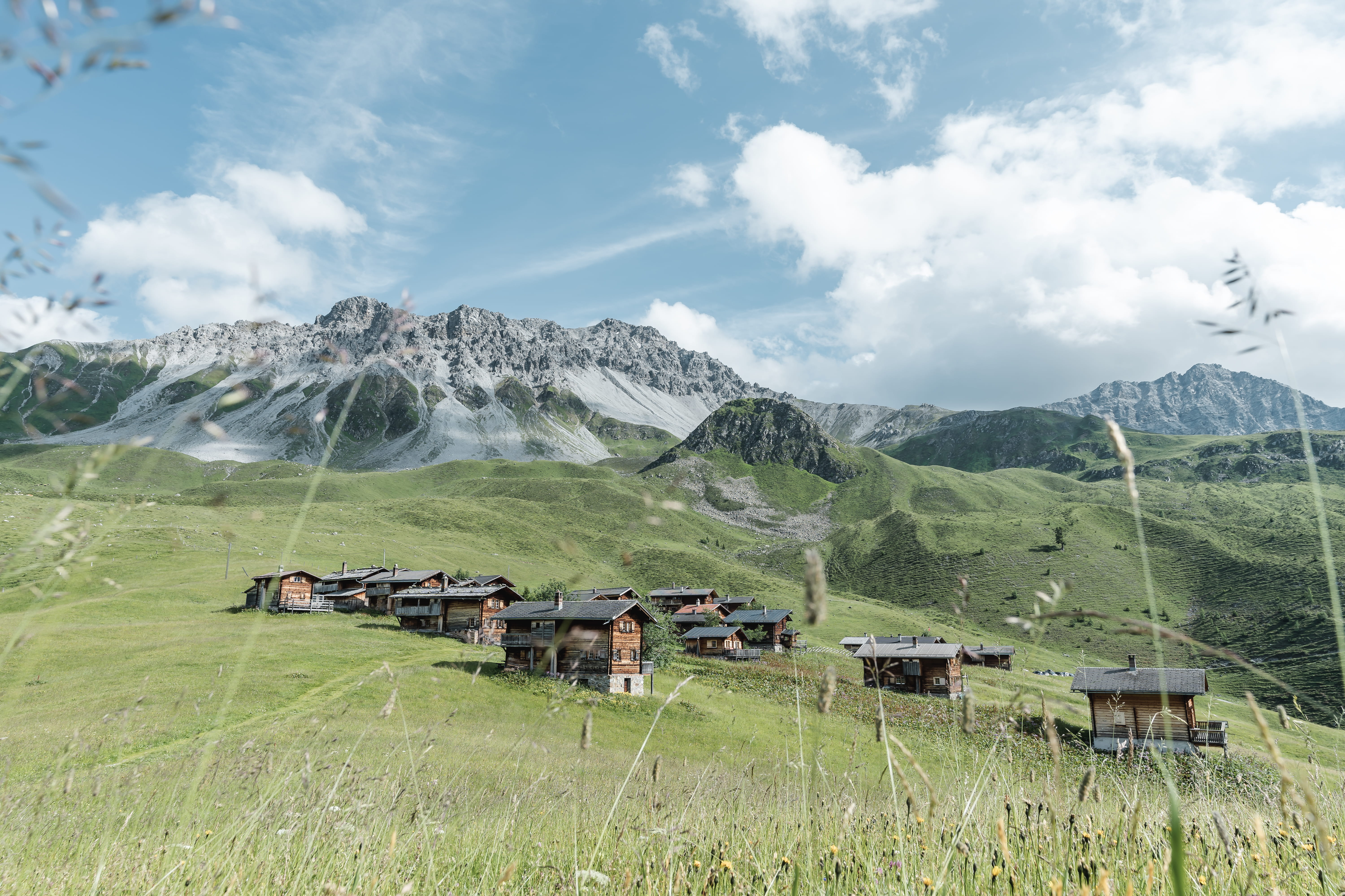 Bergdorf mit traditionellen Holzchalets auf grüner Almwiese vor imposanter Bergkulisse unter blauem Himmel mit Wolken