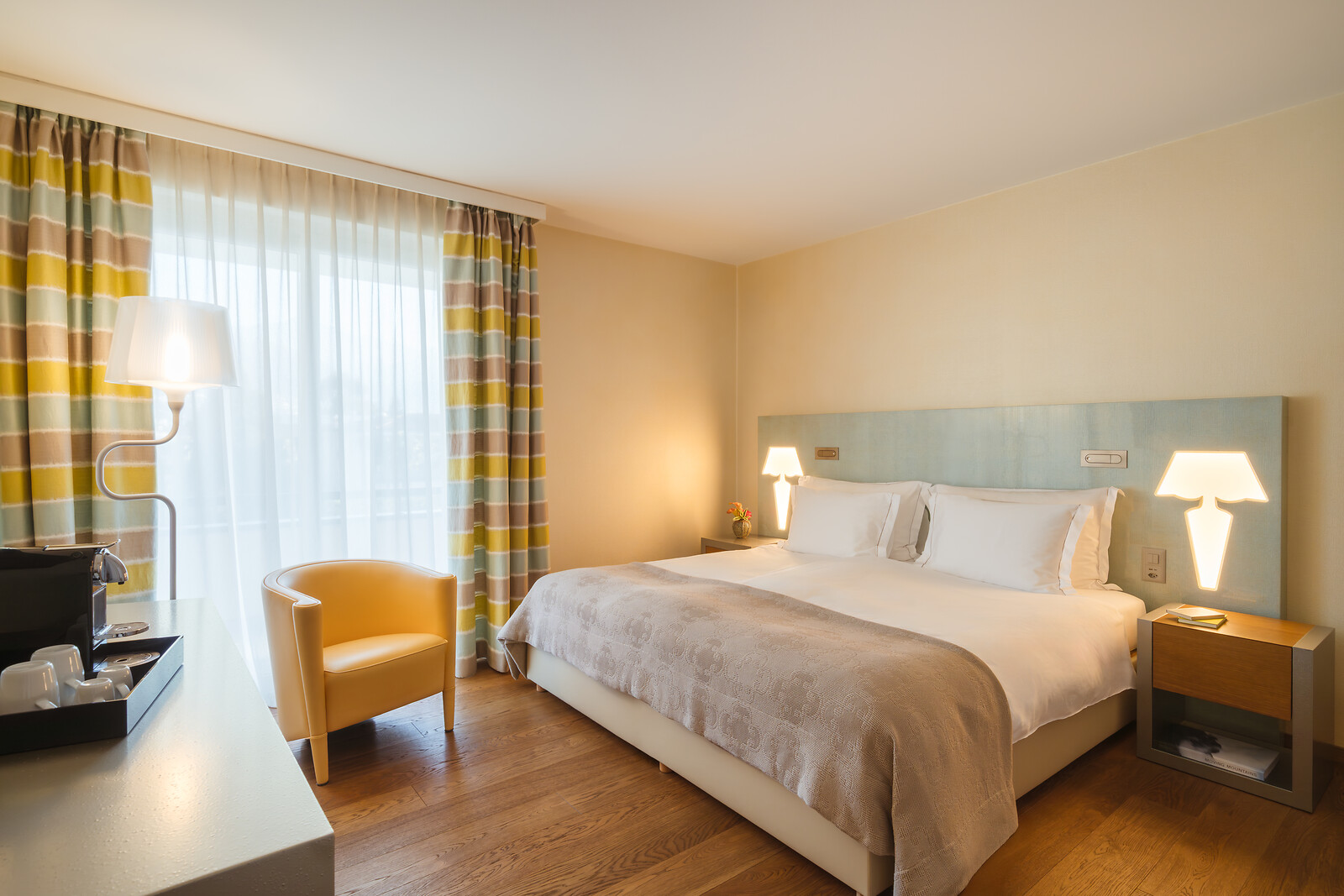 Freundliches Hotelzimmer mit großem Doppelbett, hellblauem Kopfteil, Parkettboden, gelbem Designersessel, Stehlampe und farbenfrohen Vorhängen mit Streifenmuster.
