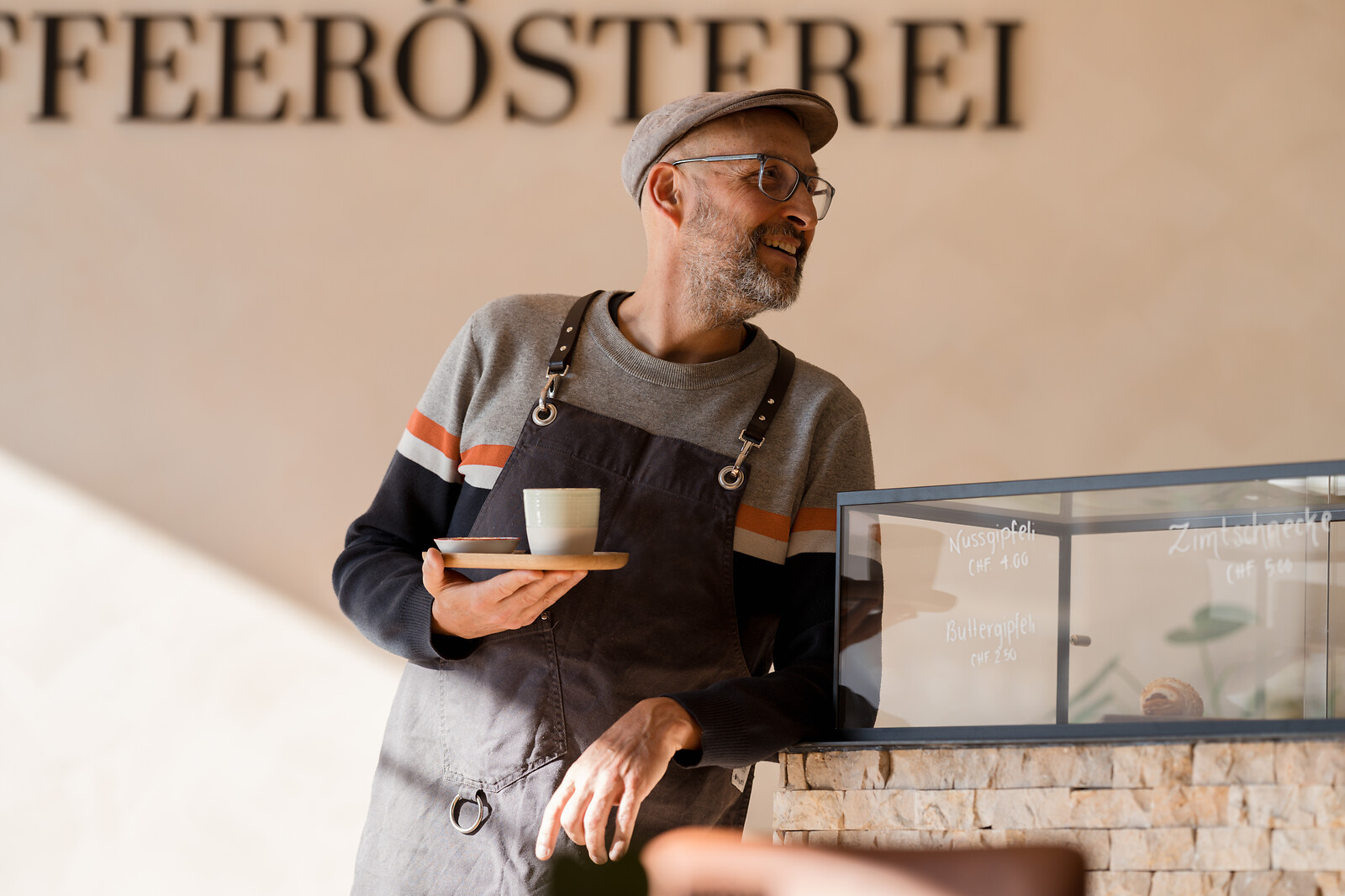 Smiling man in blue polo and cap standing with crossed arms beside shelves displaying coffee bags and Coffee Roasting.