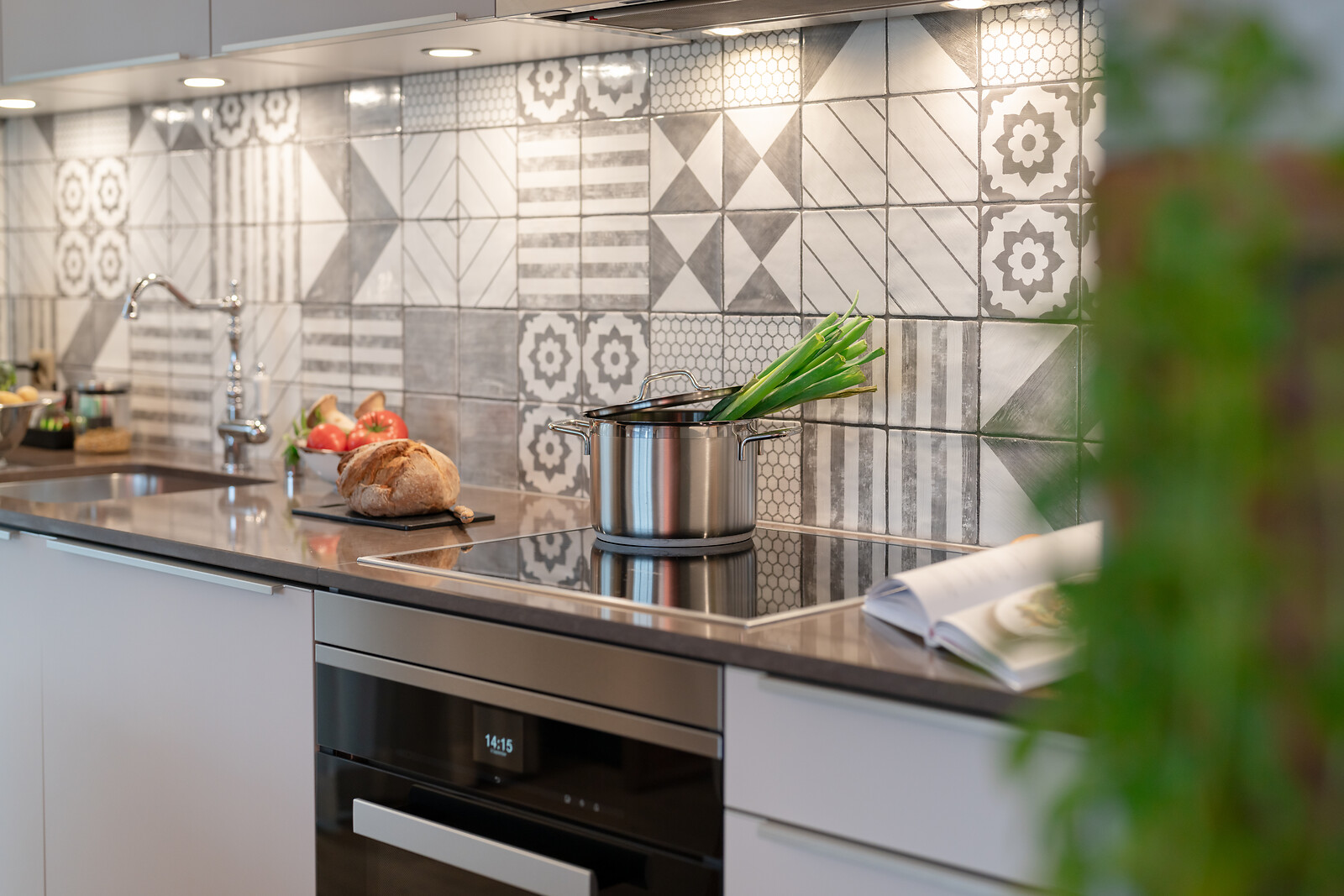 Modern kitchen area with stovetop, pot and fresh ingredients, featuring a decorative tiled backsplash and built-in appliances.