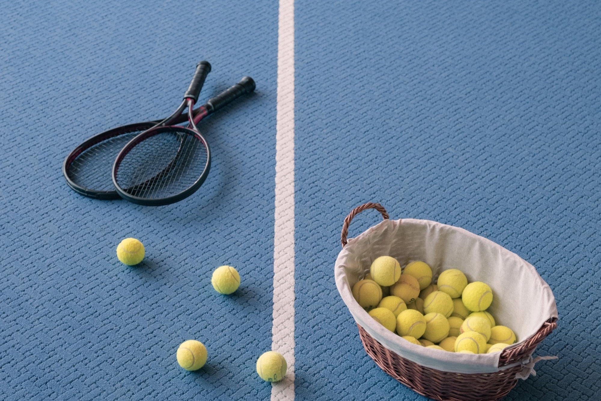 Tennis rackets and yellow balls on a blue court with a basket of balls beside the white center line.