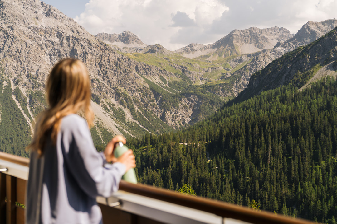 Frau auf einem Balkon mit einer Trinkflasche, die auf eine eindrucksvolle Alpenlandschaft mit felsigen Berggipfeln, grünen Hängen und dichten Wäldern unter teilweise bewölktem Himmel blickt.