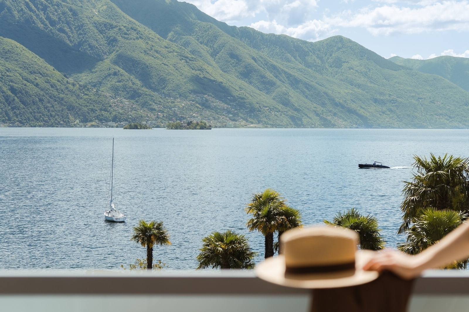 Scenic lake view with green mountains, palm trees, boats on blue water, and a sun hat in the foreground.