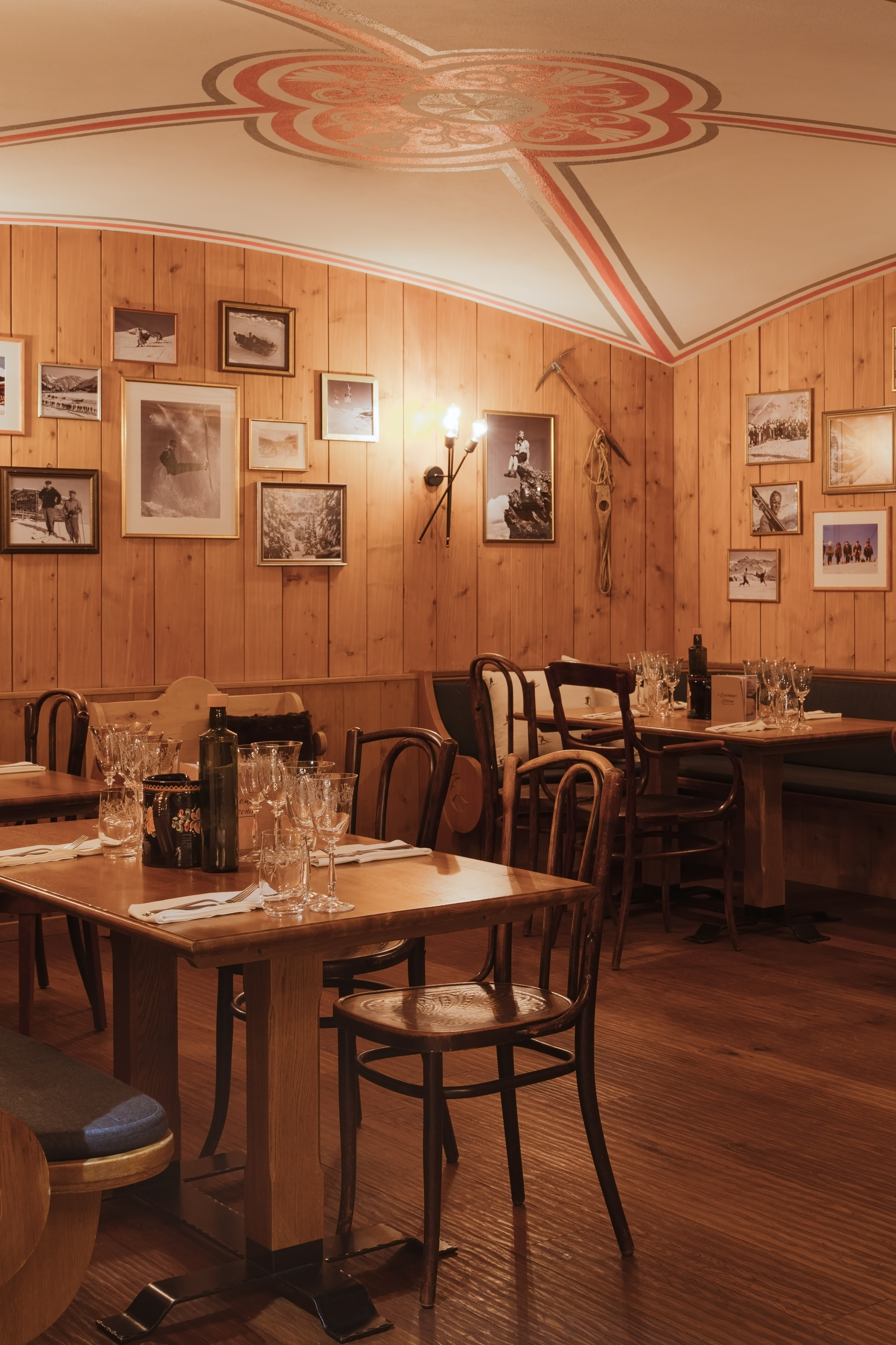 Cozy restaurant interior with wood-paneled walls, vintage ski photographs, decorative ceiling medallion, and tables set with wine glasses.