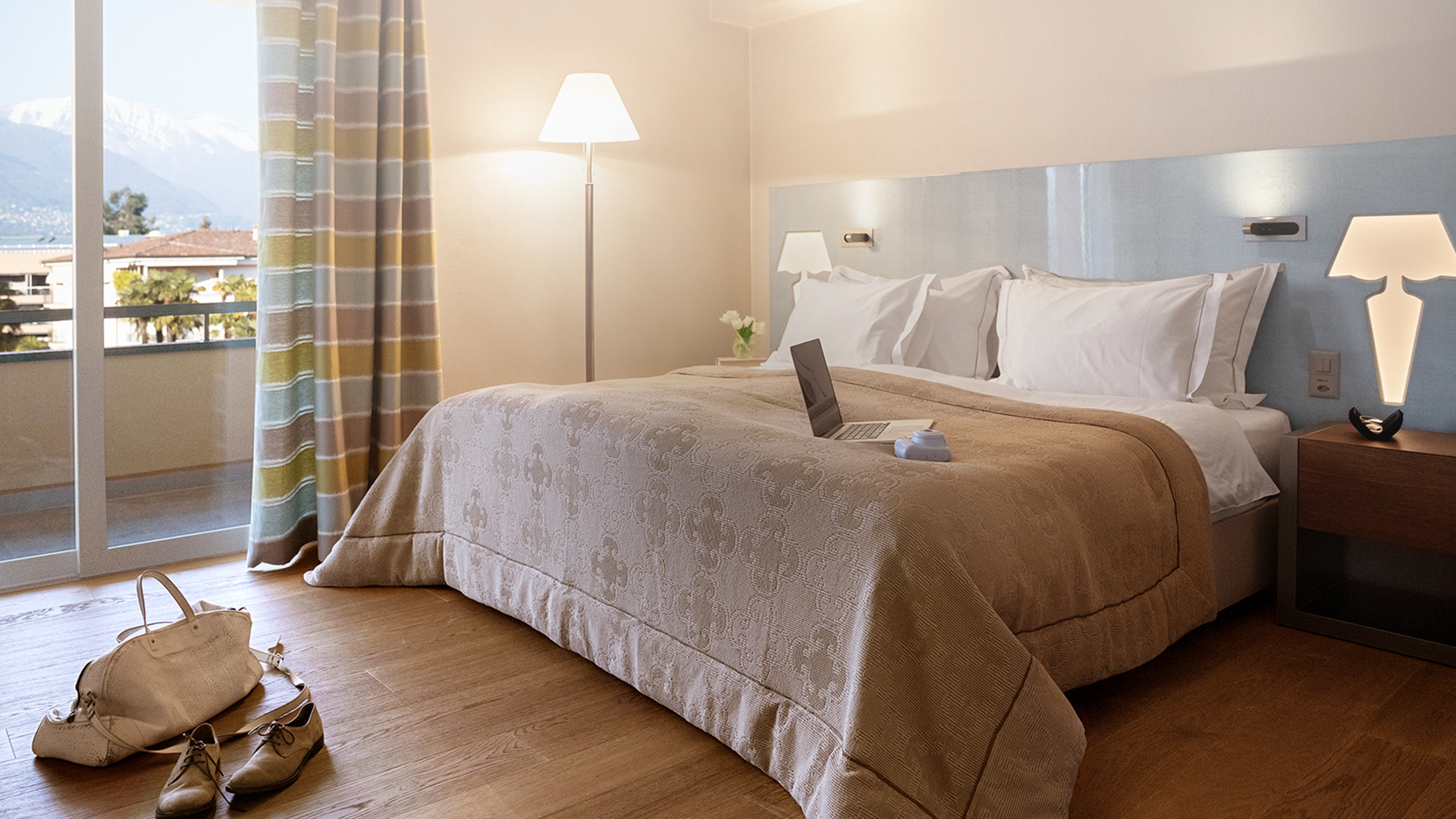 Modernes Schlafzimmer mit großem Bett, Holzboden, farbigen Blockstreifen-Vorhängen, Balkon mit Blick auf Berge und See, sowie Laptop auf dem Bett.