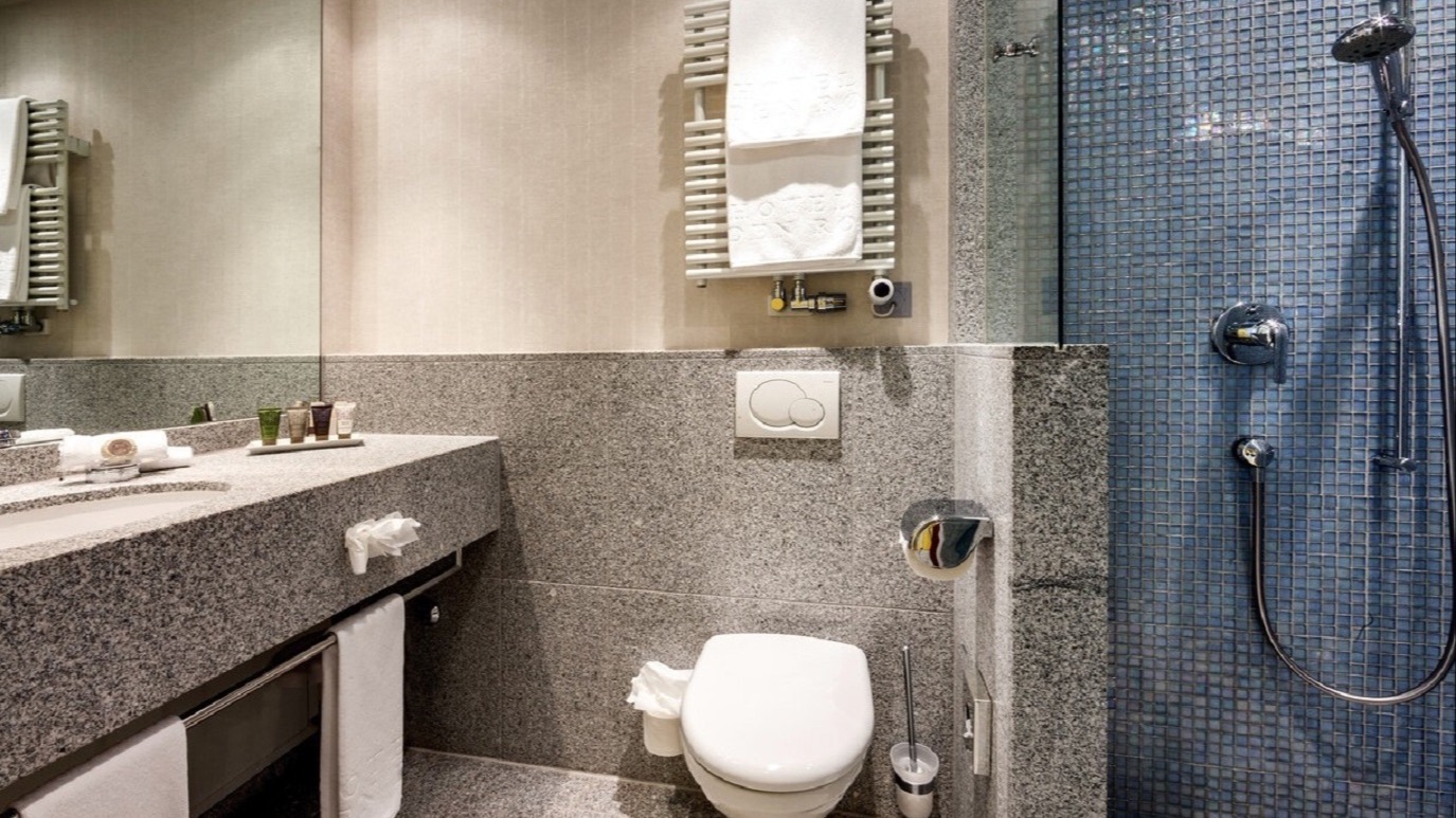 Compact hotel bathroom featuring blue mosaic-tiled shower, wall-mounted toilet, heated towel rack, and granite sink with toiletries.