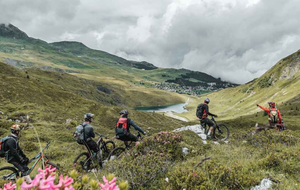 Mountain bikers with backpacks traverse a grassy alpine hillside overlooking a lake and valley under cloudy skies.