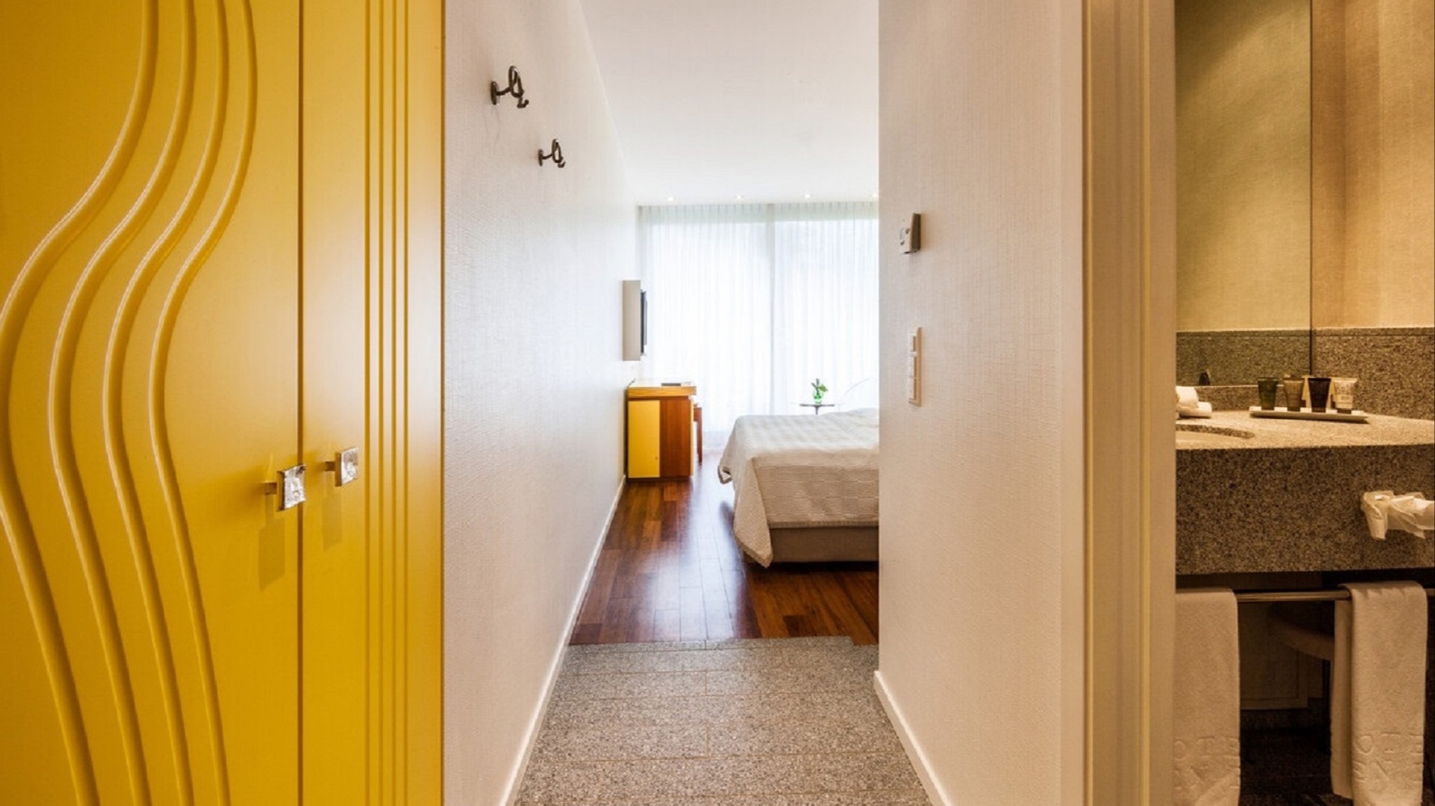 Hotelgang mit wellenförmig designtem gelbem Schrank, heller Wandfarbe, Blick ins Zimmer mit Doppelbett, Tageslicht und Zugang zum Badezimmer.