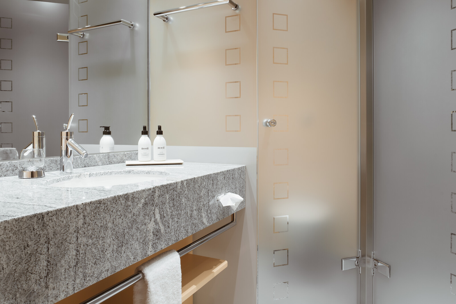 Modernes Hotelbadezimmer mit Granitwaschtisch und Waschbecken, grosser Spiegelwand, Chromarmaturen und einer Milchglas-Duschkabine mit dezenten quadratischen Mustern.