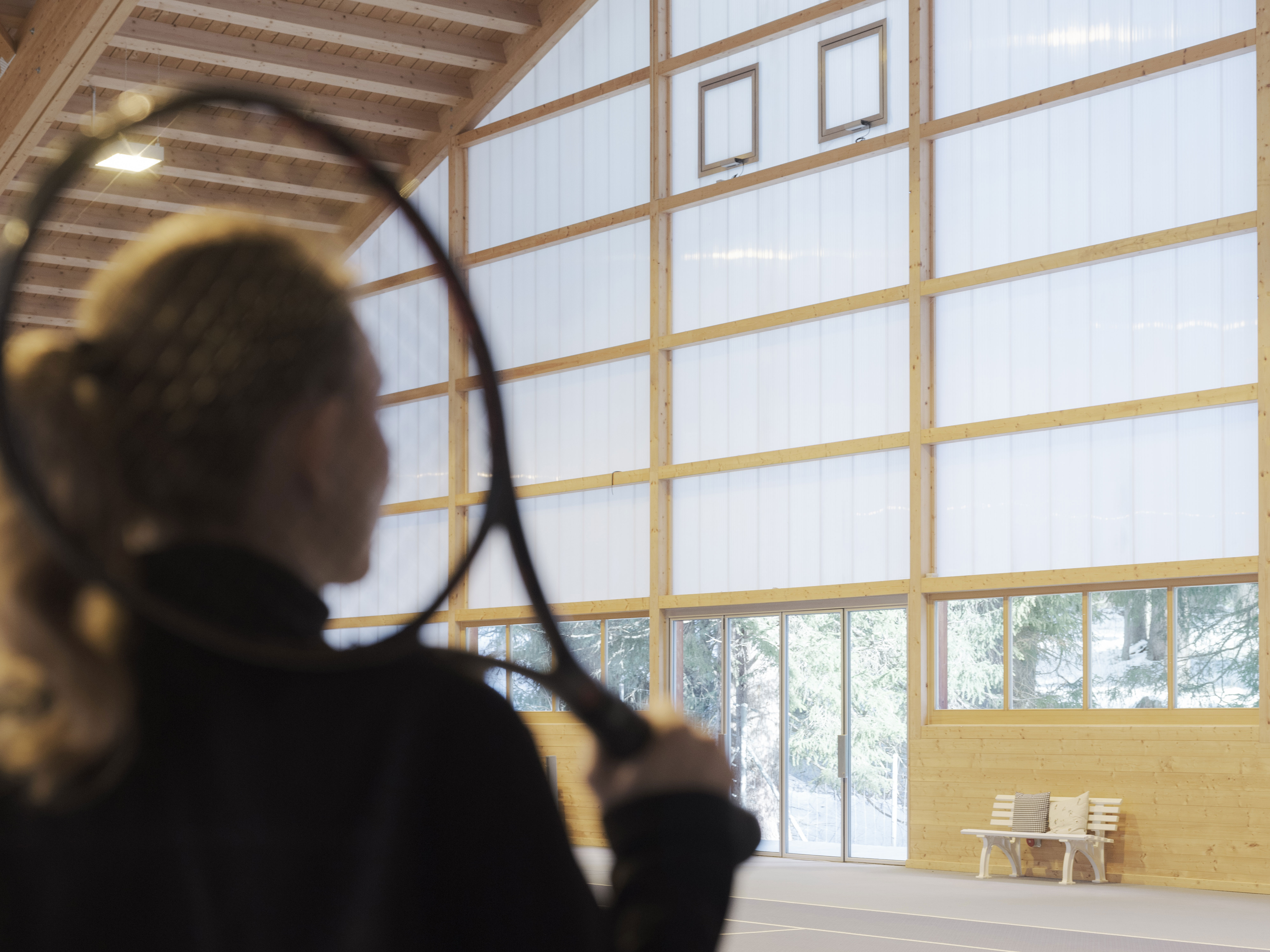 Eine Person mit Tennisschläger steht im Vordergrund einer hellen Tennishalle mit Holzkonstruktion und großen Fensterflächen.